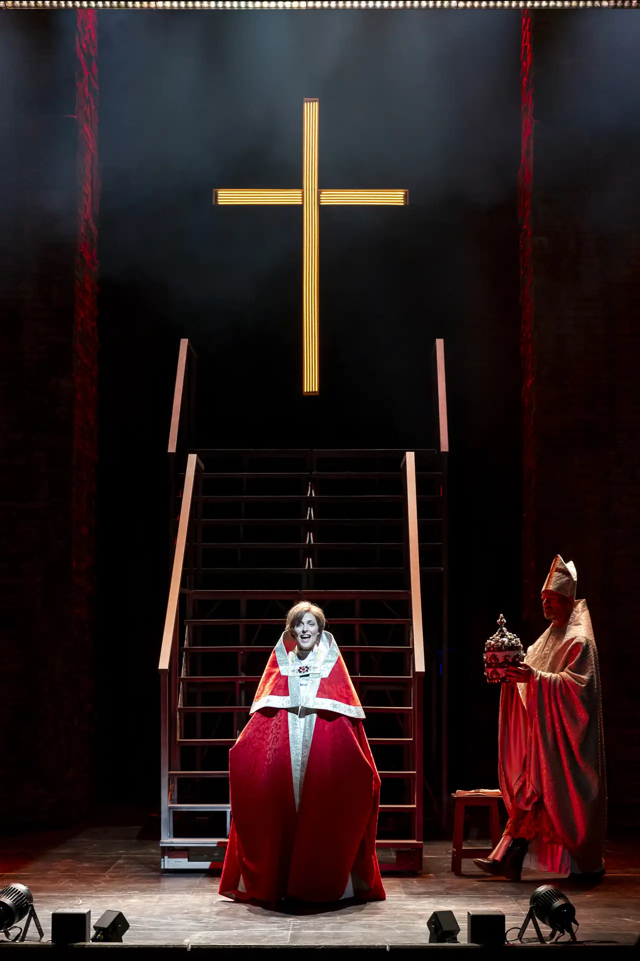 Die Päpstin – Das Musical: Person in rotem Gewand sitzt vor einer Treppe mit großem goldenem Kreuz an der Wand, daneben steht eine weitere Person in rotem Gewand mit einem Zepter.