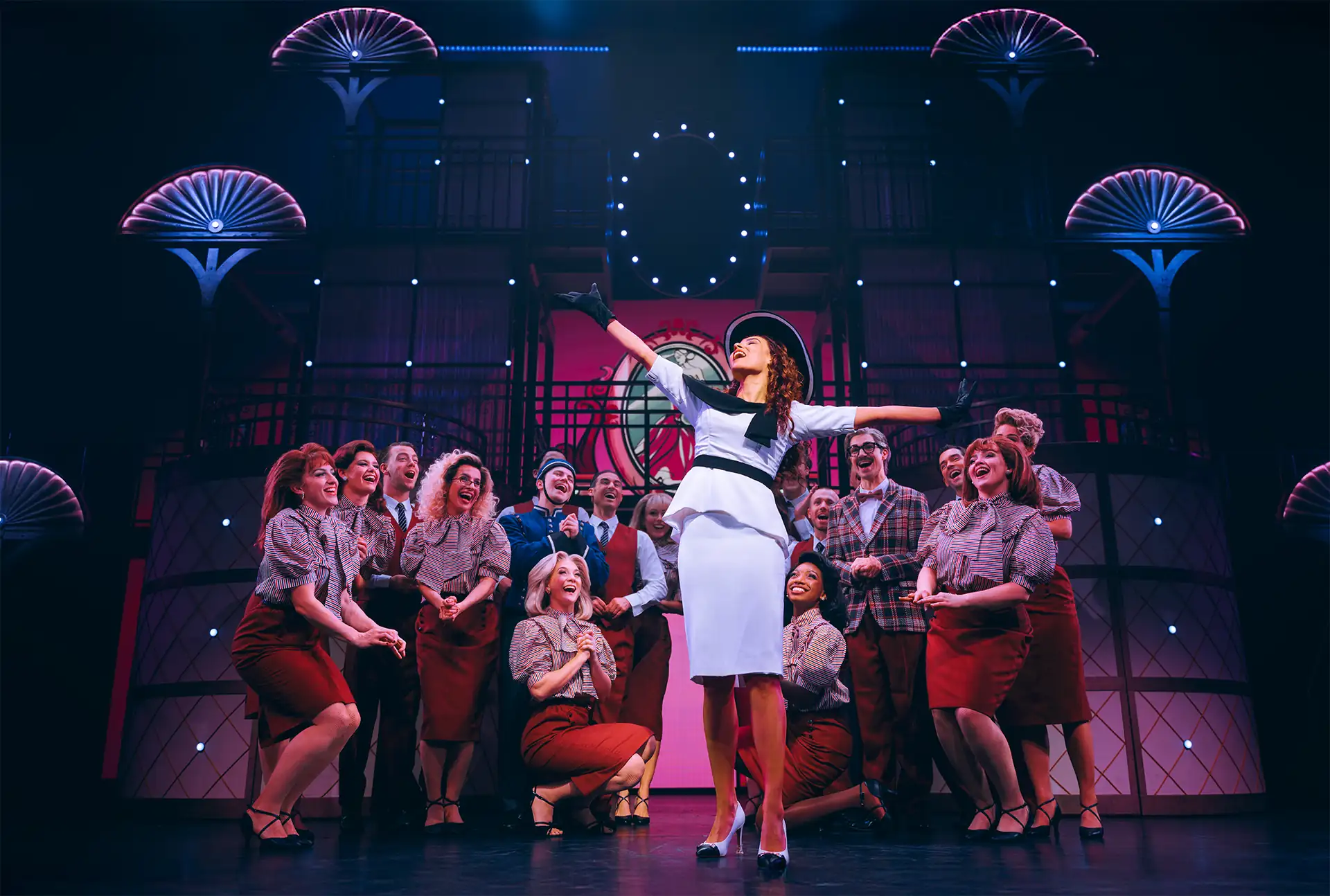 Pretty Woman Musical Szene: Eine Frau in weißem Kleid und Hut (Vivian) steht mit ausgebreiteten Armen auf einer Bühne, umgeben von mehreren Personen in karierten Hemden und roten Röcken oder Hosen, Bühnenbild mit Uhr und leuchtenden Fächern im Hintergrund.