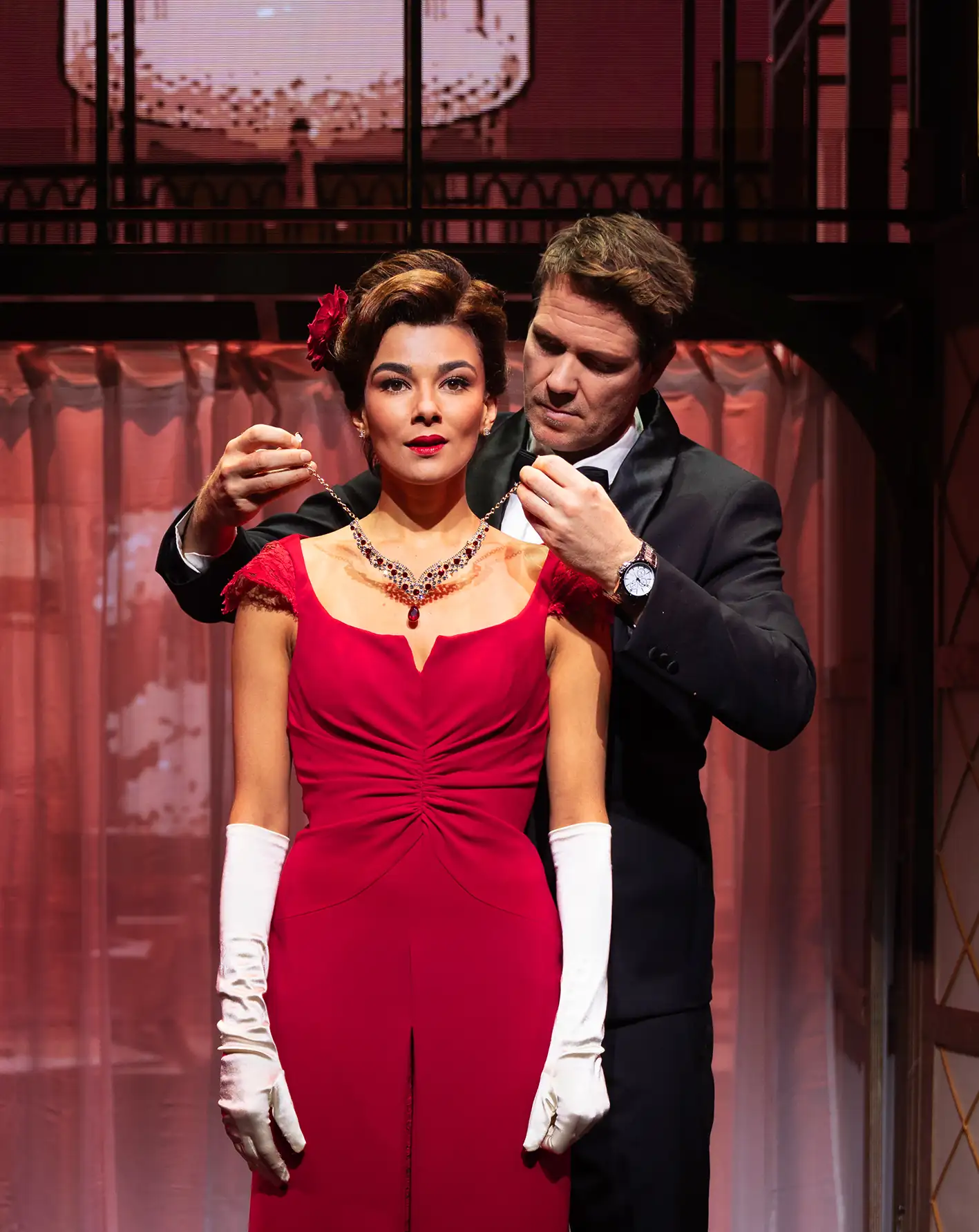 Pretty Woman Musical Szene: Eine Frau in rotem Abendkleid mit weißen langen Handschuhen (Vivian) trägt eine Halskette, ein Mann im schwarzen Anzug (Edward) legt ihr die Kette an. Bühnenhintergrund mit Vorhängen und Kronleuchter.