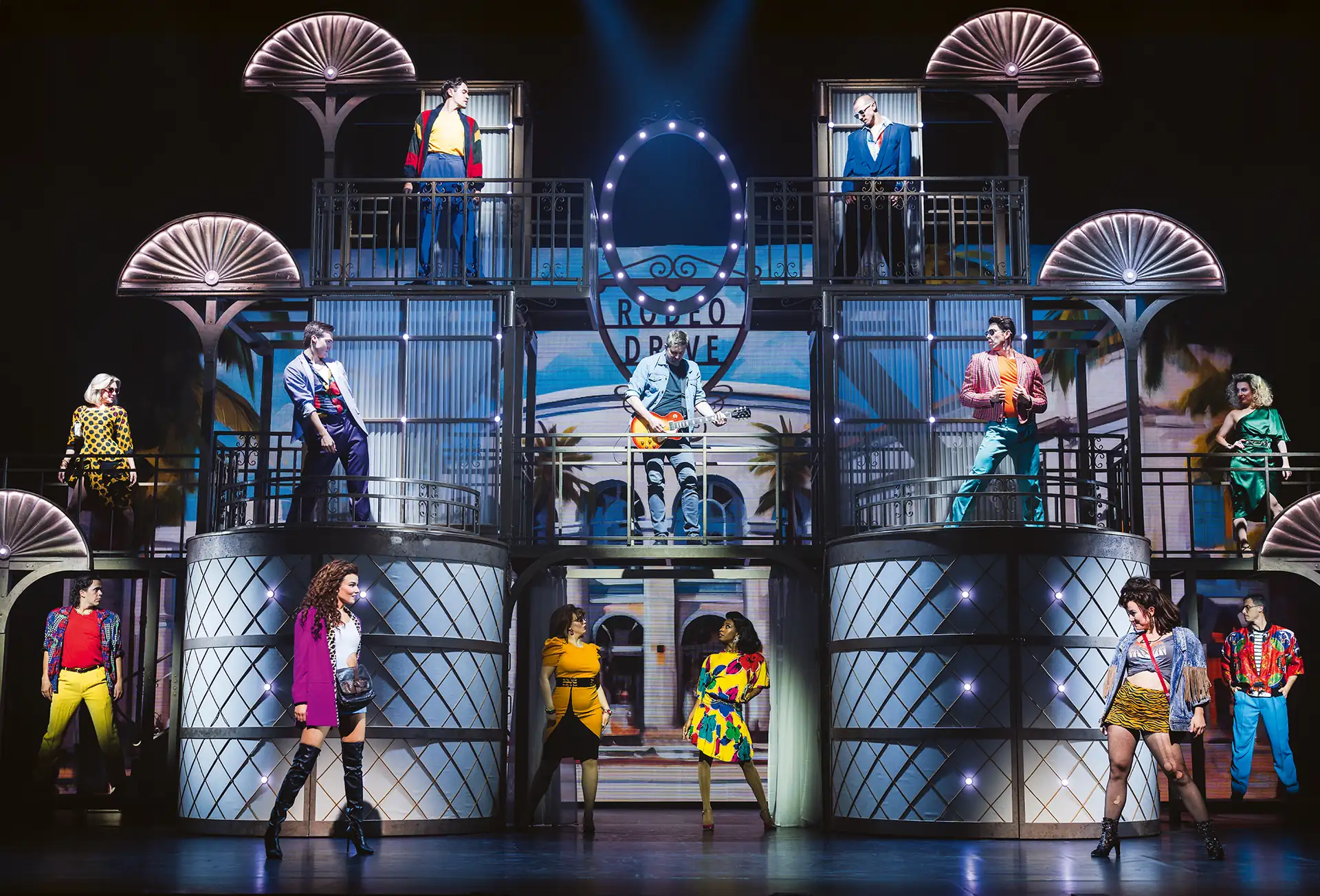 Pretty Woman Musical Szene: Bühnenbild des Pretty Woman Musicals mit mehreren Schauspielern auf zwei Ebenen vor einem Kulissenaufbau mit Neonzeichen 'RODEO DRIVE'.