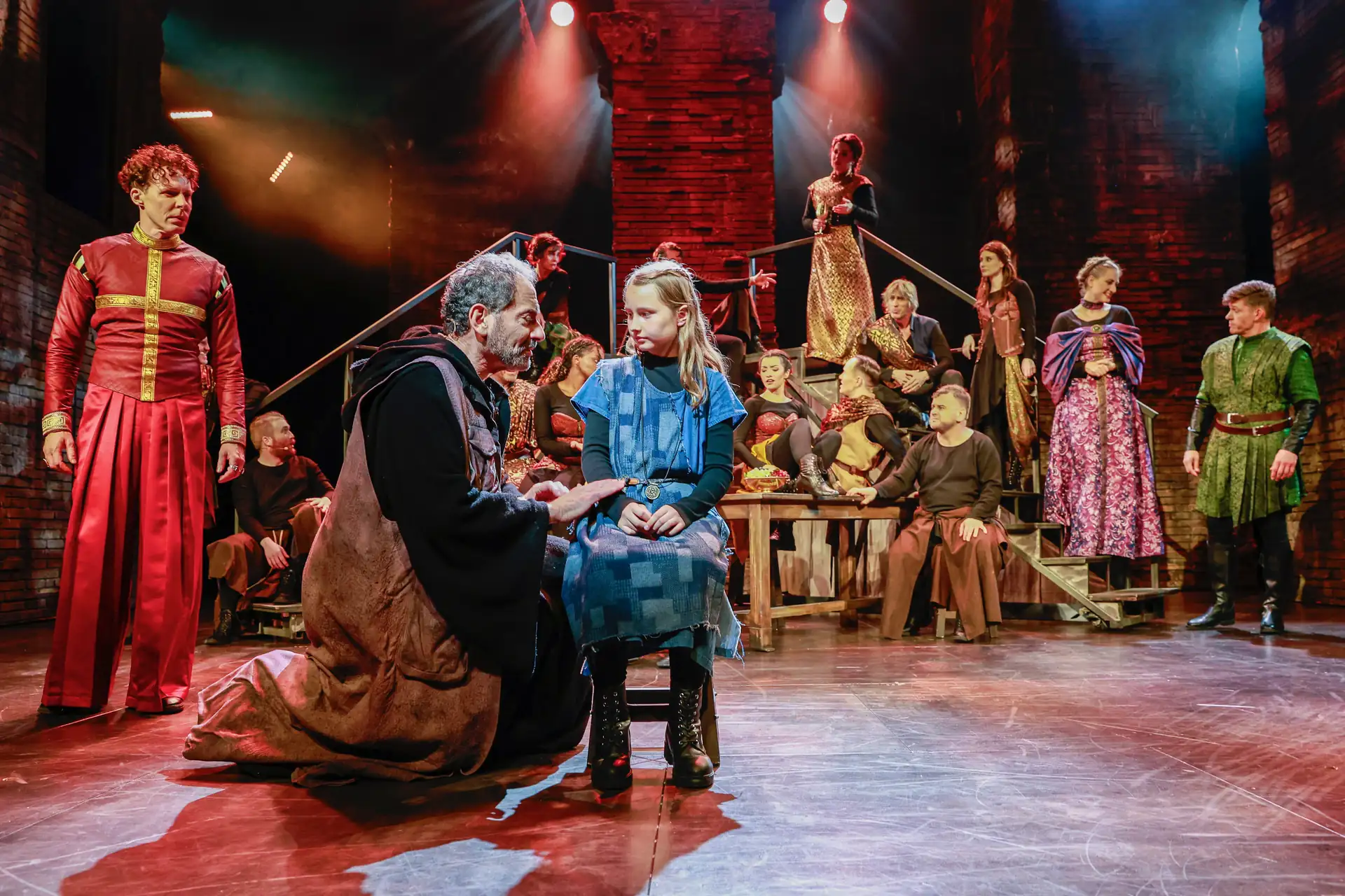 Die Päpstin – Das Musical: Szene mit mehreren Darstellern in historischen Kostümen, ein Mann kniet vor einem Mädchen auf einem Stuhl, im Hintergrund weitere Schauspieler auf einer Bühne mit rotem Backstein und Scheinwerferlicht.