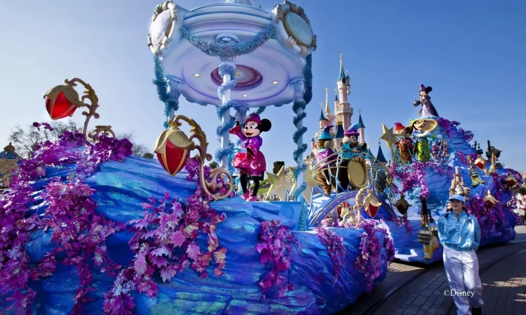 Die farbenfrohe „Parade on Main Street“ im Disneyland Paris zeigt Minnie Mouse, die auf einem prächtigen, blau-violetten Wagen steht, umgeben von leuchtenden Dekorationen und fantasievollen Elementen. Im Hintergrund ragen das ikonische Schloss und weitere Disney-Figuren auf Wagen empor. Die Parade versprüht festliche Energie, während Darsteller in passenden Kostümen die Route entlanglaufen und die Menge begeistert. Minnie Mouse trägt ein elegantes Outfit und lächelt den Zuschauern zu, während sie auf ihrem glitzernden Wagen durch den Park fährt.