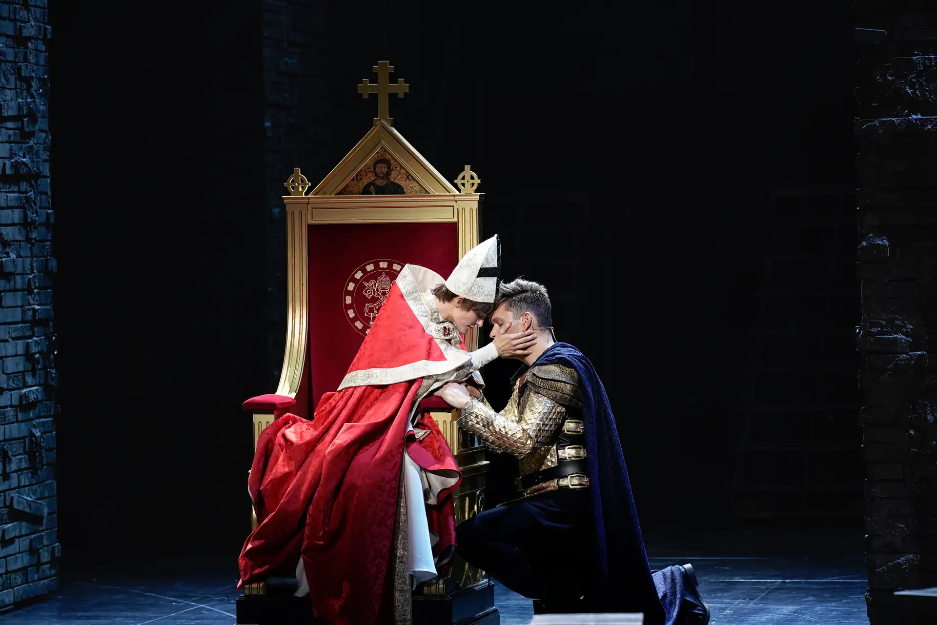 Die Päpstin – Das Musical: Eine Person in rotem Gewand und weißer Mitra beugt sich zu einer knienden Person in goldener Rüstung und blauem Umhang vor einem goldenen Thron mit Kreuz und Ikone vor.