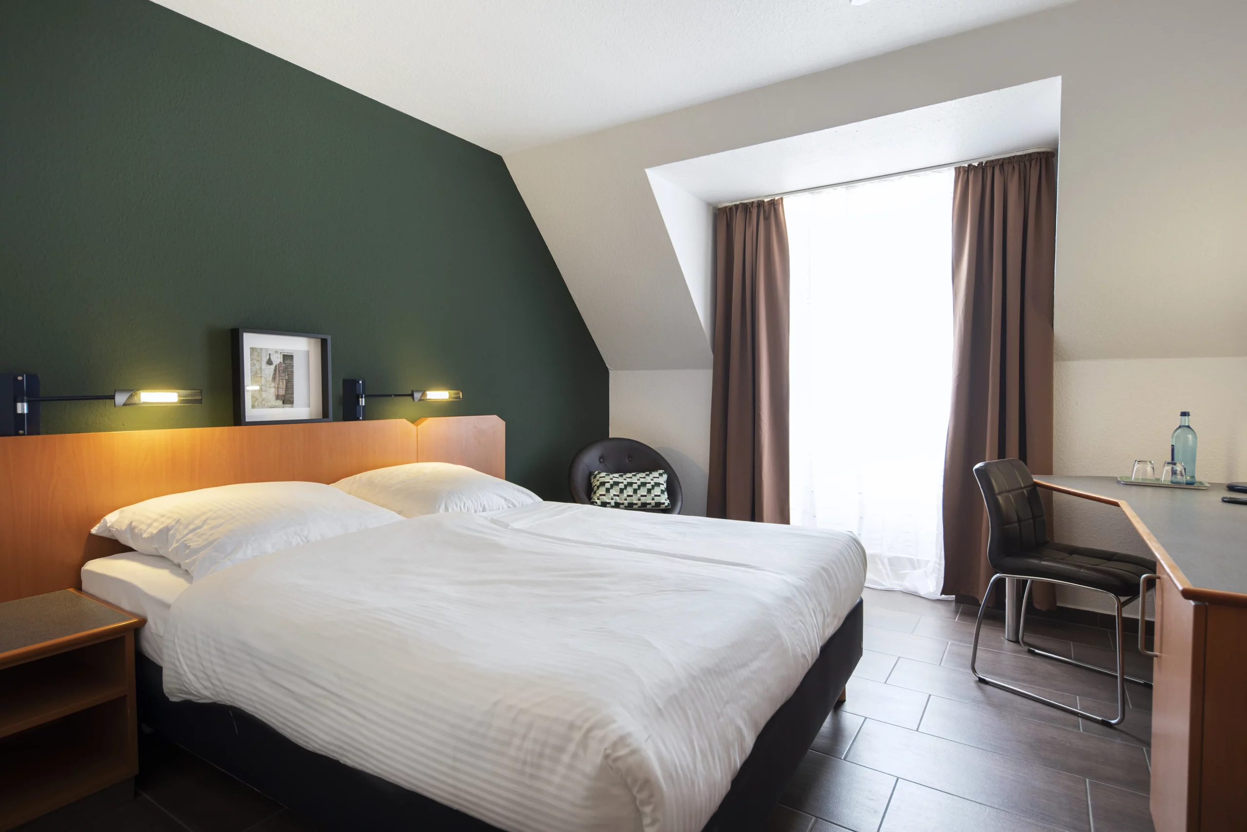 Hotelzimmer mit Doppelbett vor dunkelgrüner Wand, Nachttisch, Sessel neben Fenster mit Vorhängen und Schreibtisch mit Stuhl und Wasserflasche. Im ACHAT Hotel Heppenheim