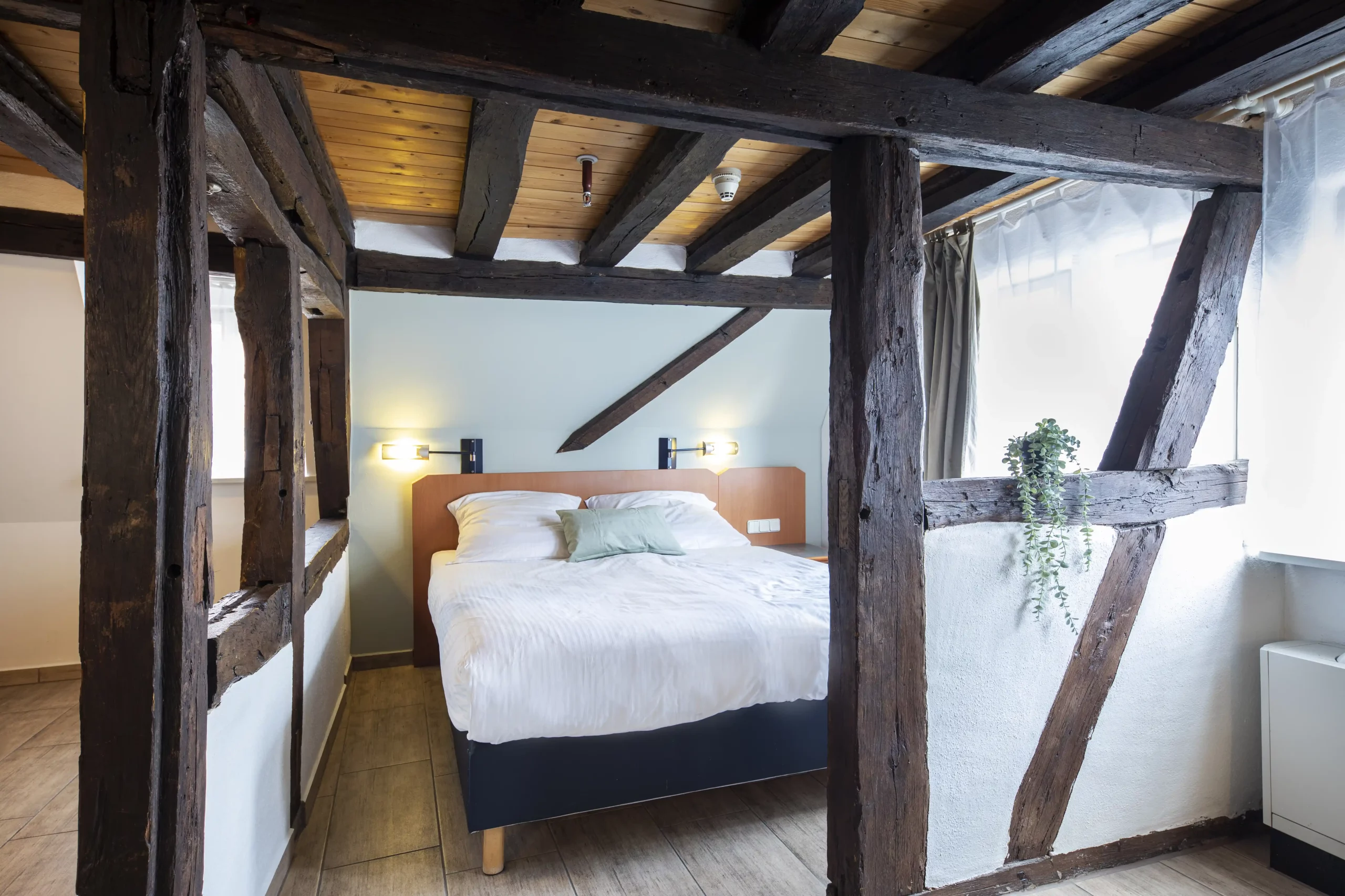 Schlafzimmer mit sichtbaren dunklen Fachwerkbalken, Doppelbett mit weißer Bettwäsche und zwei Wandlampen an der weißen Wand im ACHAT Hotel Heppenheim