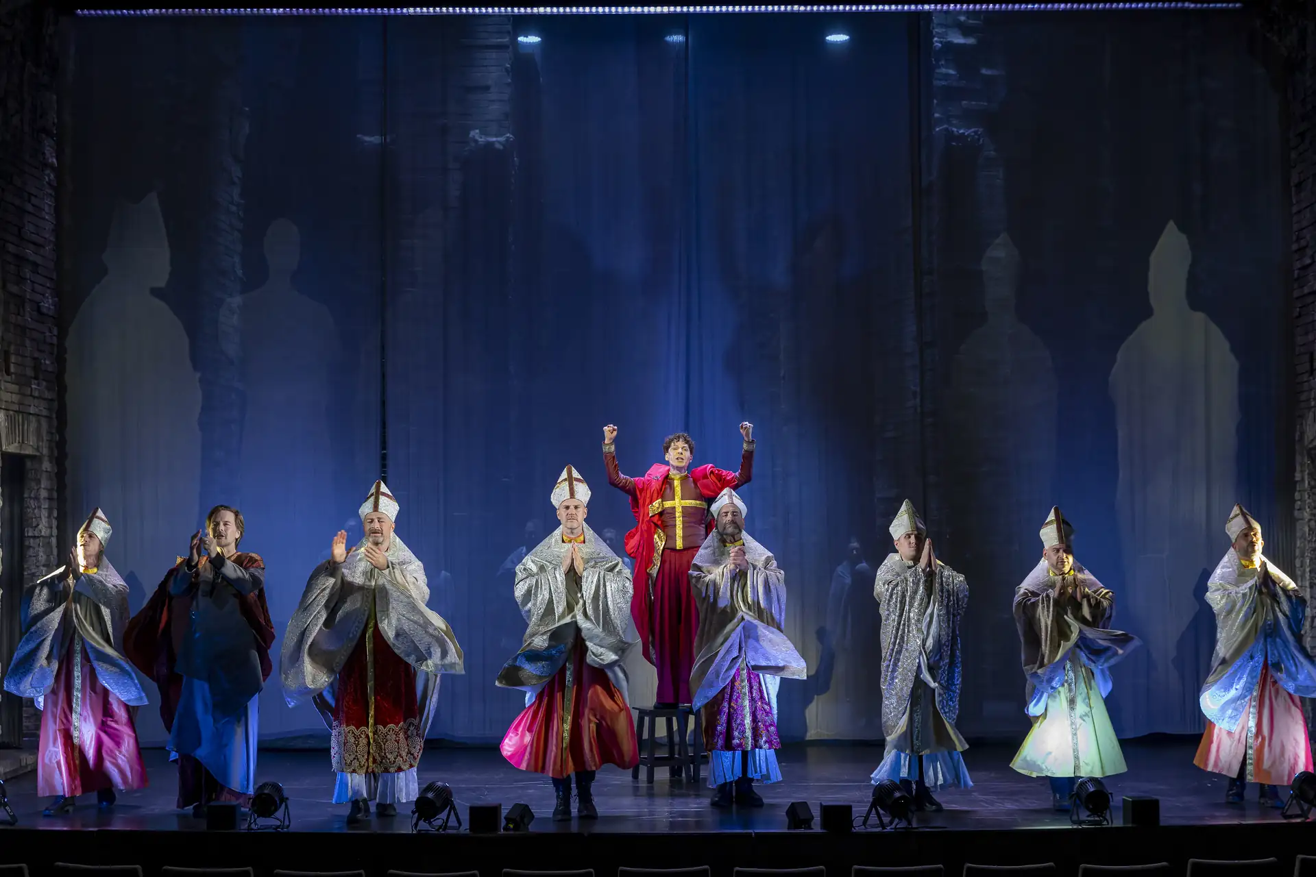 Die Päpstin – Das Musical: Acht Personen in historischen Kostümen stehen auf einer Bühne, eine Person in der Mitte trägt ein rotes Gewand und hebt die Arme, die anderen tragen weiße Kopfbedeckungen und bunte Gewänder.
