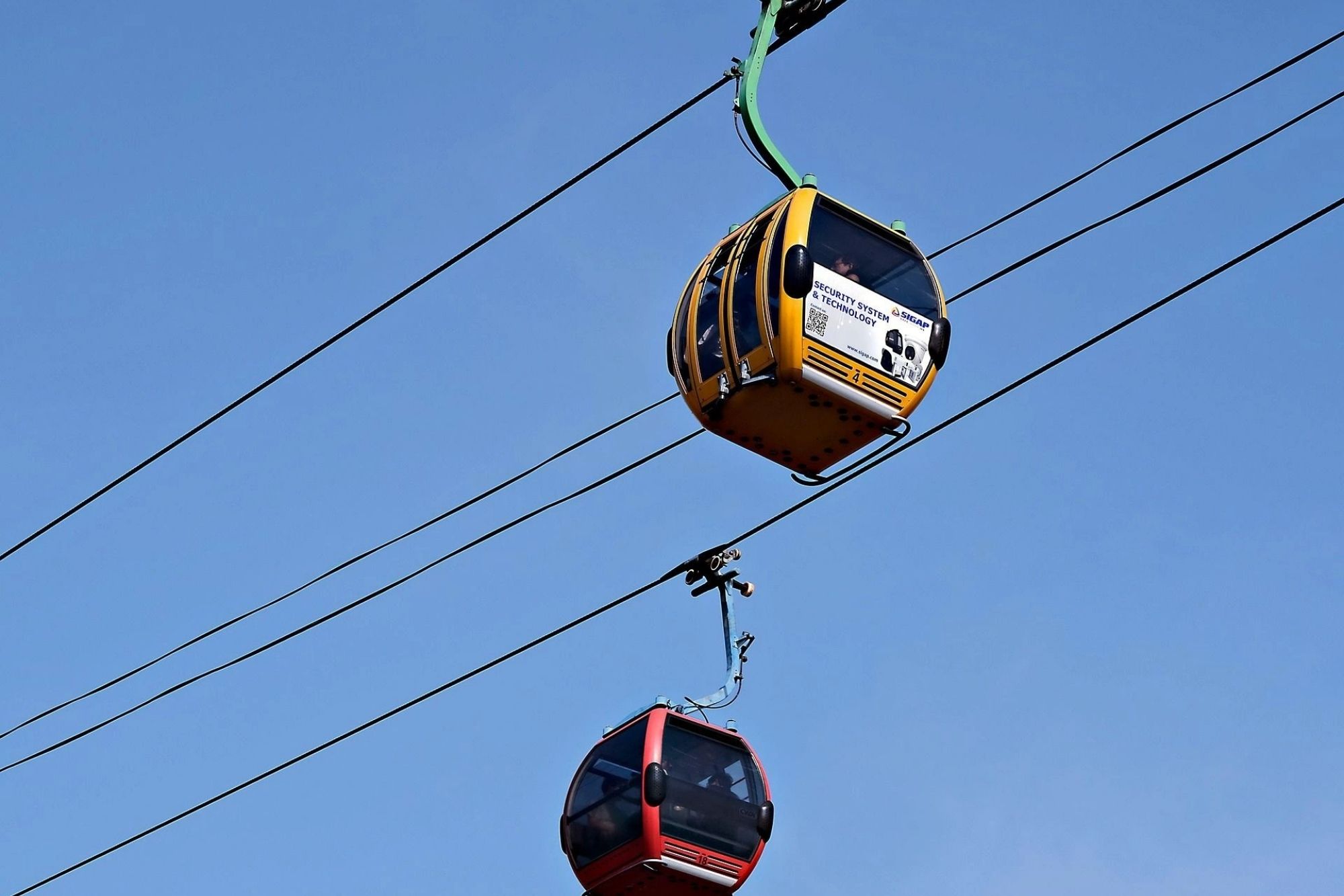 Zwei Gondelbahnkabinen, eine gelb und eine rot, fahren an Seilen vor klarem blauem Himmel