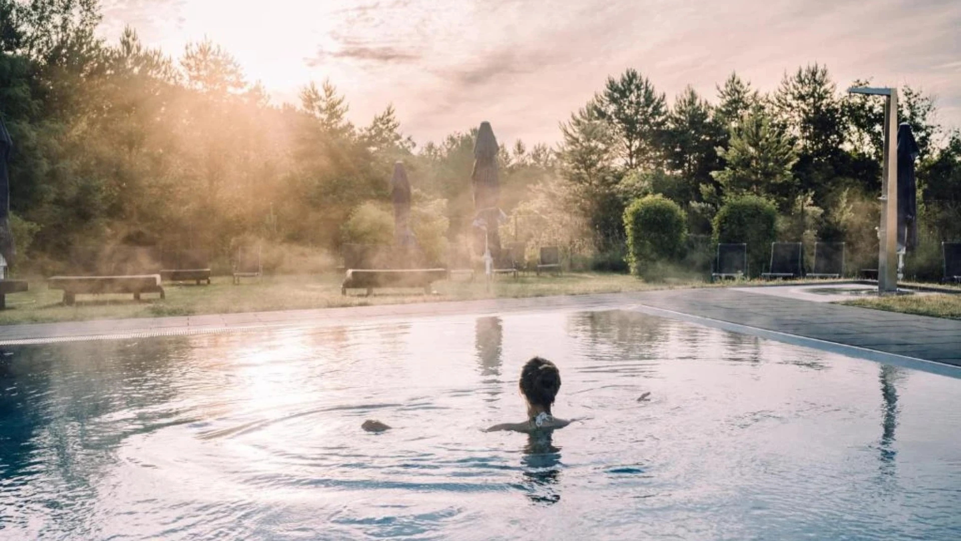 Ein idyllischer Außenpool im aja Resort Bad Saarow: Umgeben von grüner Natur und blauen Sonnenschirmen lädt der Pool mit glitzerndem Wasser zu einem entspannten Start in den Tag ein. Die sanften Sonnenstrahlen am Morgen sorgen für eine ruhige und erfrischende Atmosphäre.