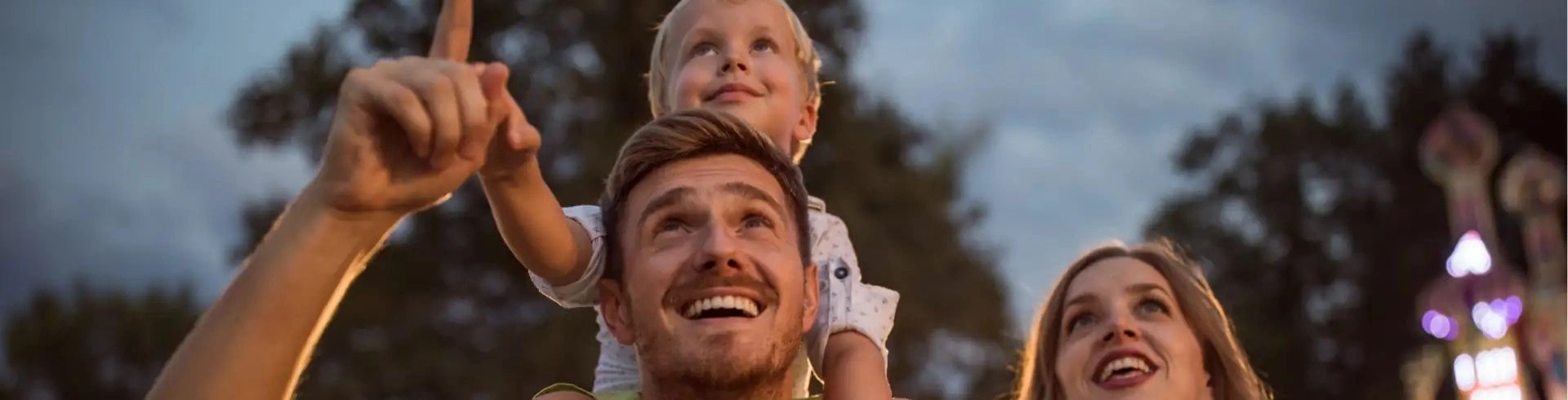 Event- und Erlebnisreisen Symbolbild im Bannerformat: Vater mit Kind auf den Schultern und Mutter daneben blicken staunend hoch, im Hintergrund sind verschwommene Lichter einer Open Air-Veranstaltung.