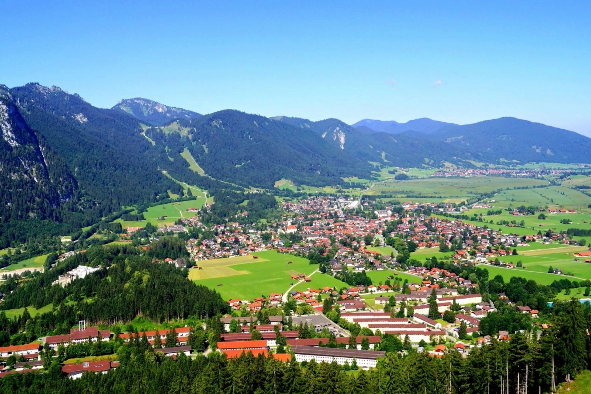 Blick auf Tal und Oberammergau, umgeben von bewaldeten Bergen und grünen Feldern unter blauem Himmel