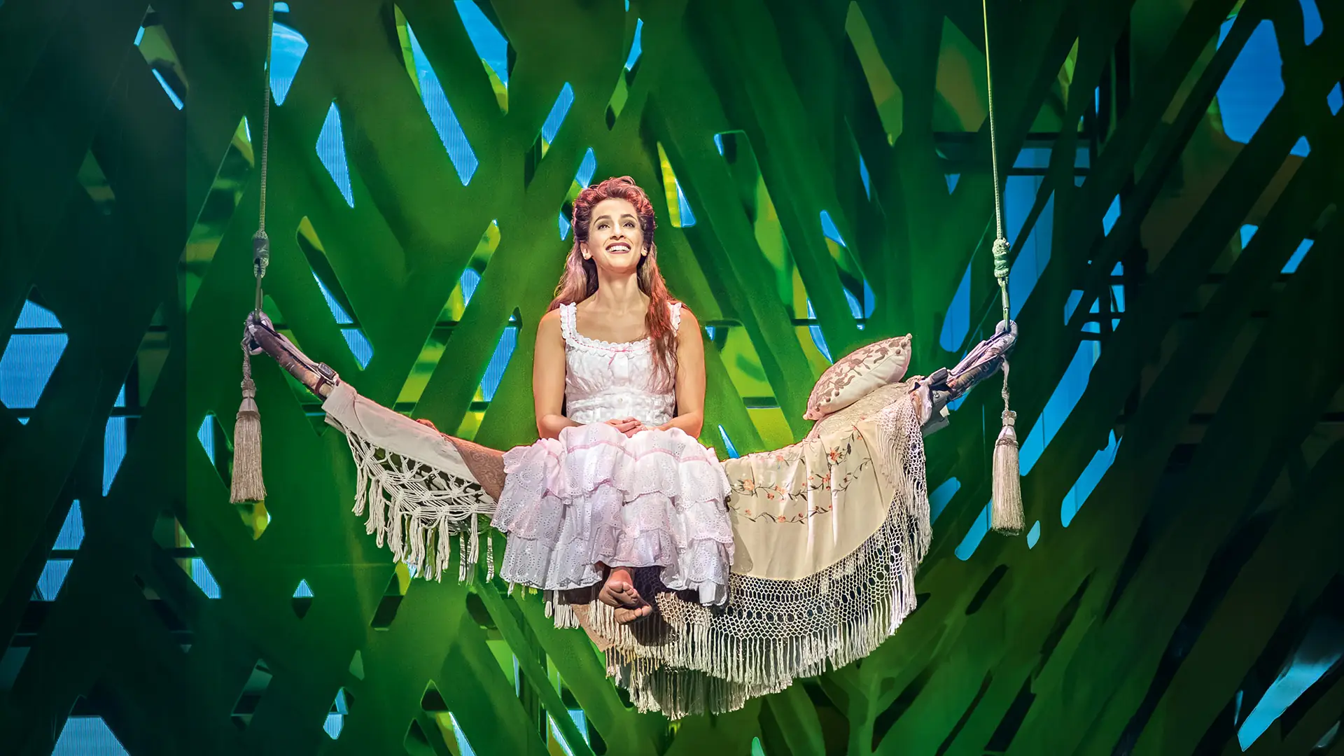 Szenenbild aus dem Musical Tarzan 2025. Eine Frau in weißem, mehrlagigem Kleid sitzt barfuß auf einer hängenden, mit Decken und Kissen bedeckten Schaukel vor grün-blauem Dschungelhintergrund.