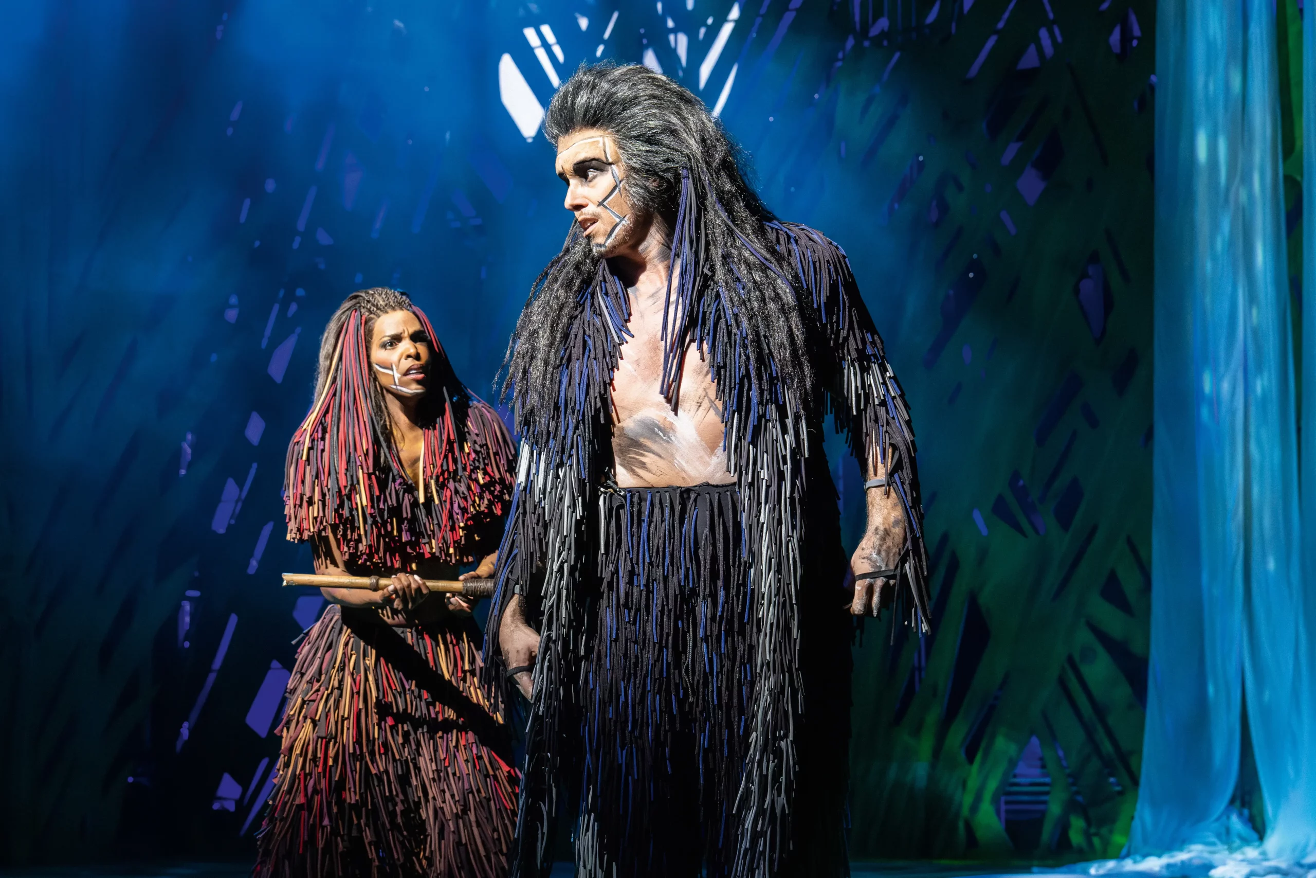 Szenenbild aus dem Musical Tarzan 2025. Zwei Darsteller in Kostümen aus langen, geflochtenen Stoffstreifen, einer mit blau-schwarzen, der andere mit rot-braunen Streifen, auf einer Bühne mit blau-grünem Hintergrund.