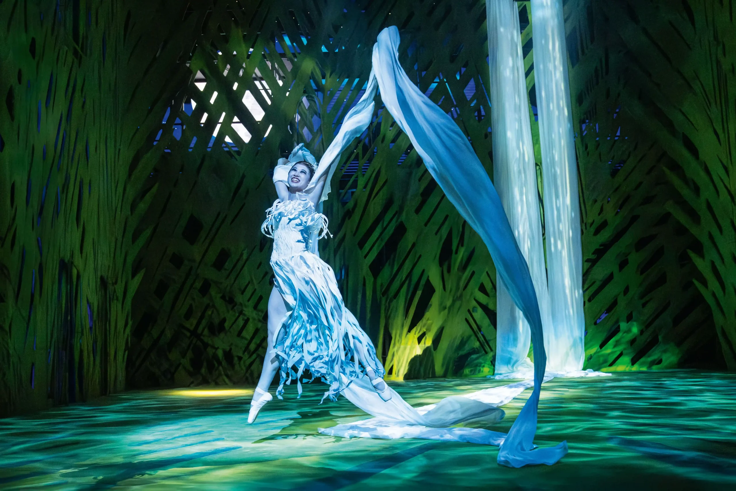 Szenenbild aus dem Musical Tarzan 2025. Tänzerin in fließendem, weißem Kostüm mit langen Stoffbahnen, die sie in der Luft schwingt, vor grün-blau beleuchtetem Bühnenhintergrund.