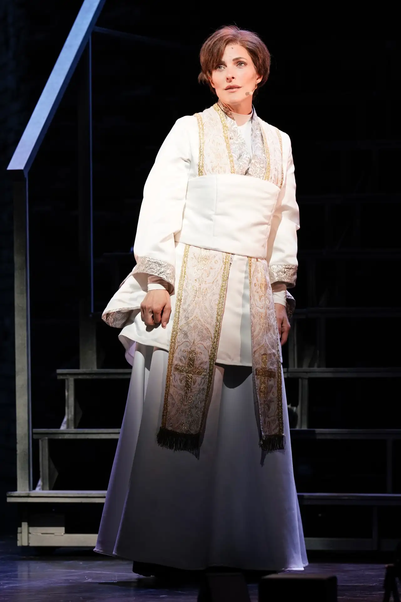 Die Päpstin – Das Musical: Person in weißem, langem Gewand mit goldverziertem liturgischem Stola auf dunkler Bühne vor Treppe.