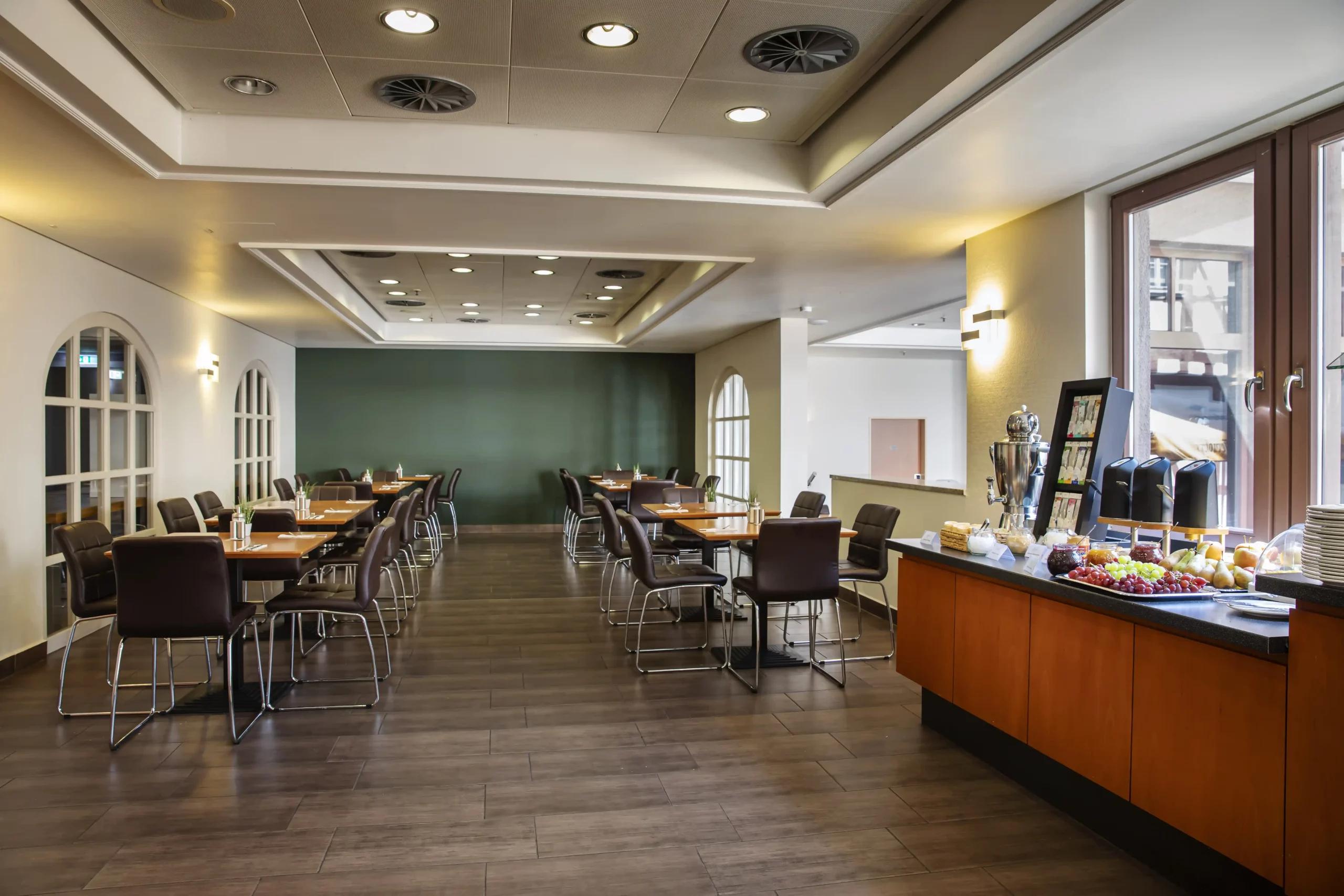 Moderner Frühstücksraum im ACHAT Hotel Heppenheim mit mehreren Tischen und Stühlen sowie einem Buffet mit Obst und Getränken am Fenster
