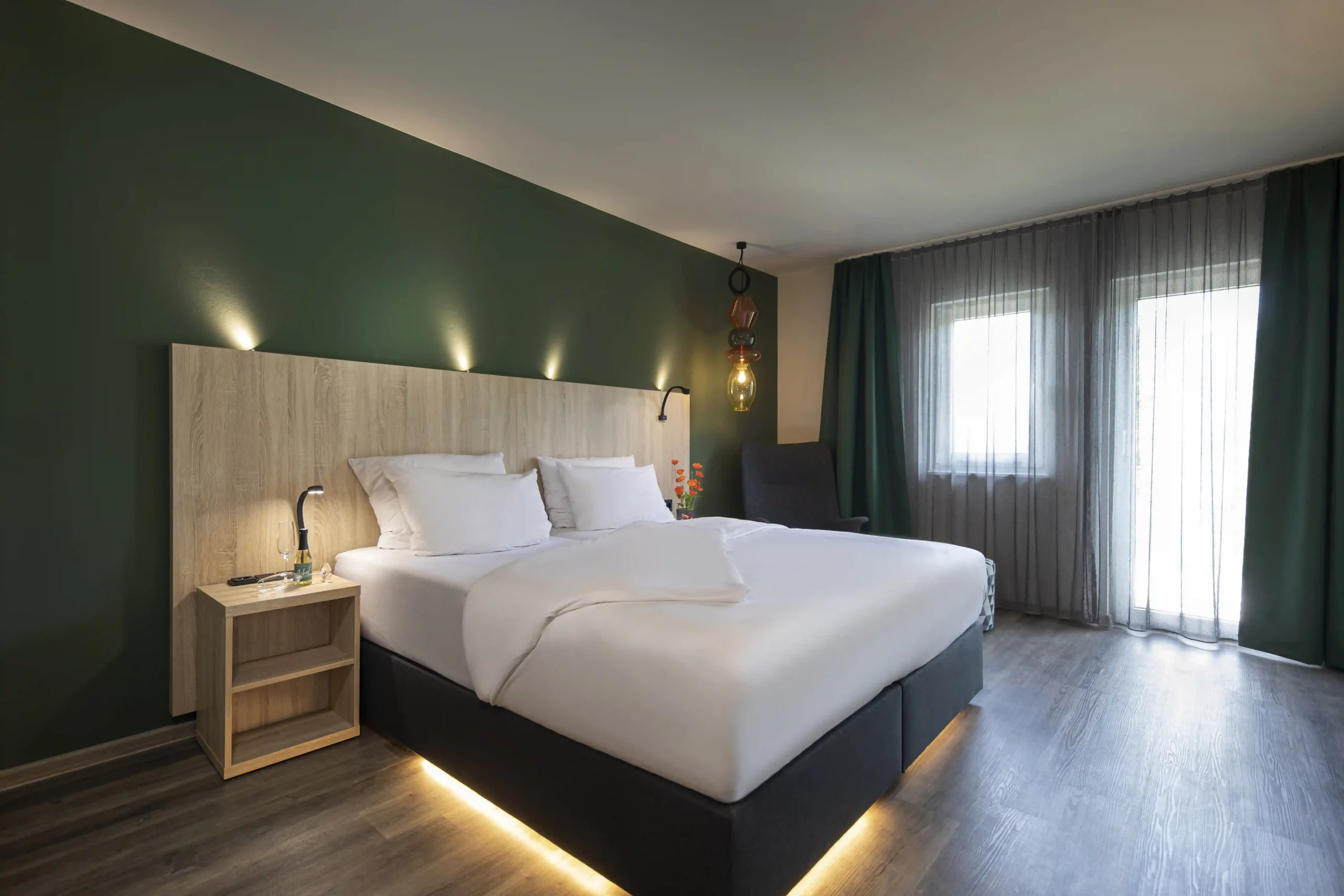 Modern eingerichtetes Hotelzimmer mit großem Doppelbett, Holz-Nachttisch, indirekter Beleuchtung unter dem Bett, grün gestrichener Wand und bodentiefen Fenstern mit halbtransparenten Vorhängen
