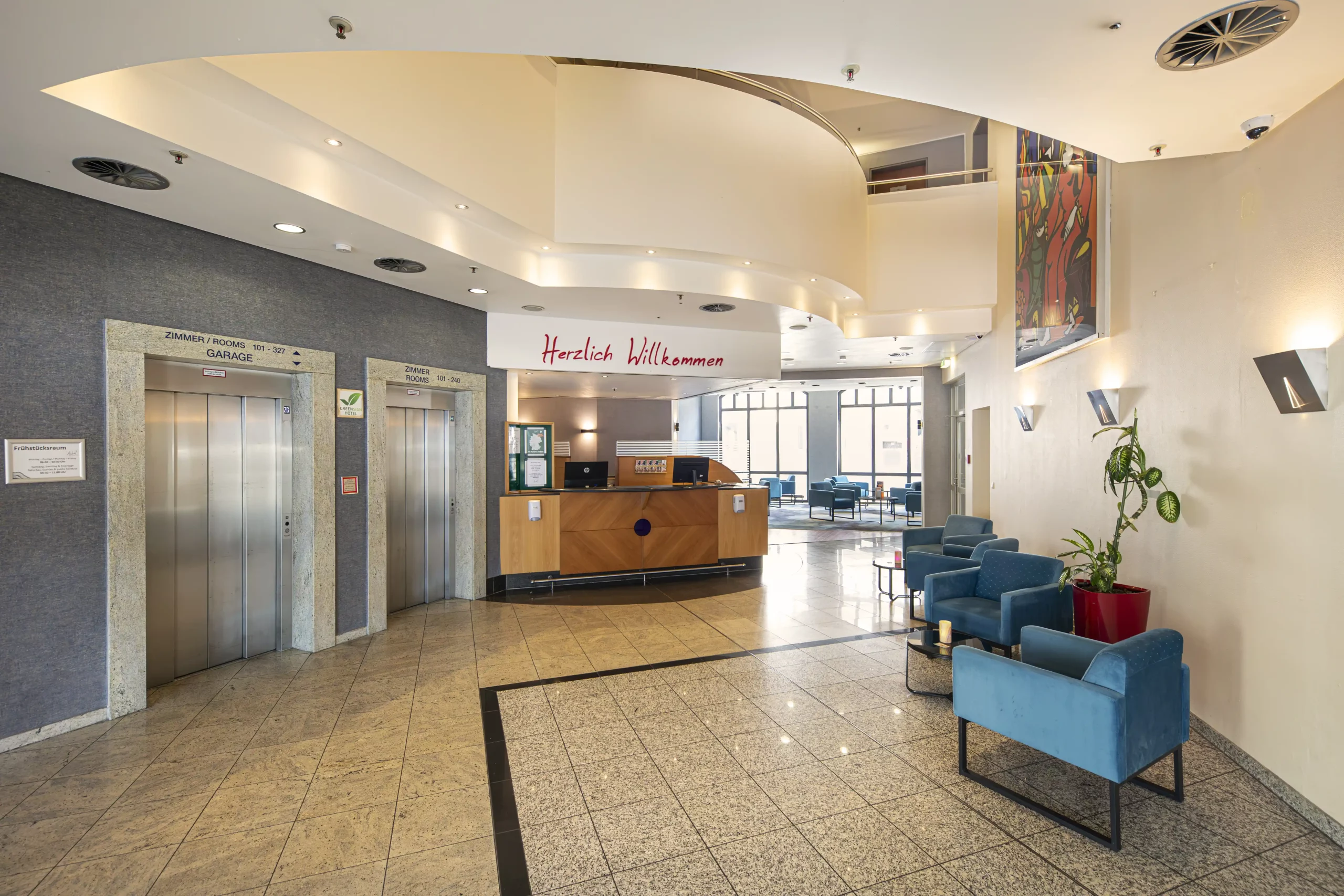 Helle Hotellobby im ACHAT Hotel Heppenheim mit Rezeption, zwei Aufzügen links, blauen Sesseln rechts und großem Fenster im Hintergrund