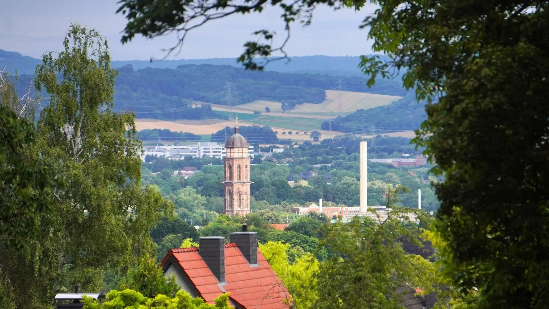 Blick auf Göttingen mit dem markanten Kirchturm im Vordergrund, umgeben von Bäumen und grünen Feldern im Hintergrund.