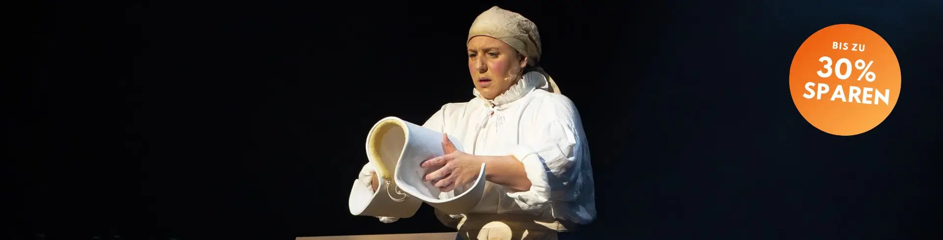 Schauspielerin von die Amme in historischer Kleidung mit Kopftuch gießt aus einem weißen Krug auf dunklem Bühnenhintergrund