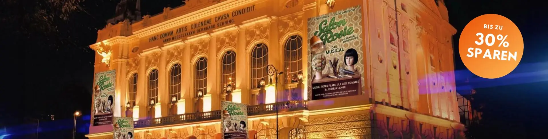 Beleuchtetes historisches Theatergebäude mit Bannern für das Musical 'Salon Rosie' und einem orangefarbenen Kreis mit der Aufschrift 'BIS ZU 30% SPAREN'