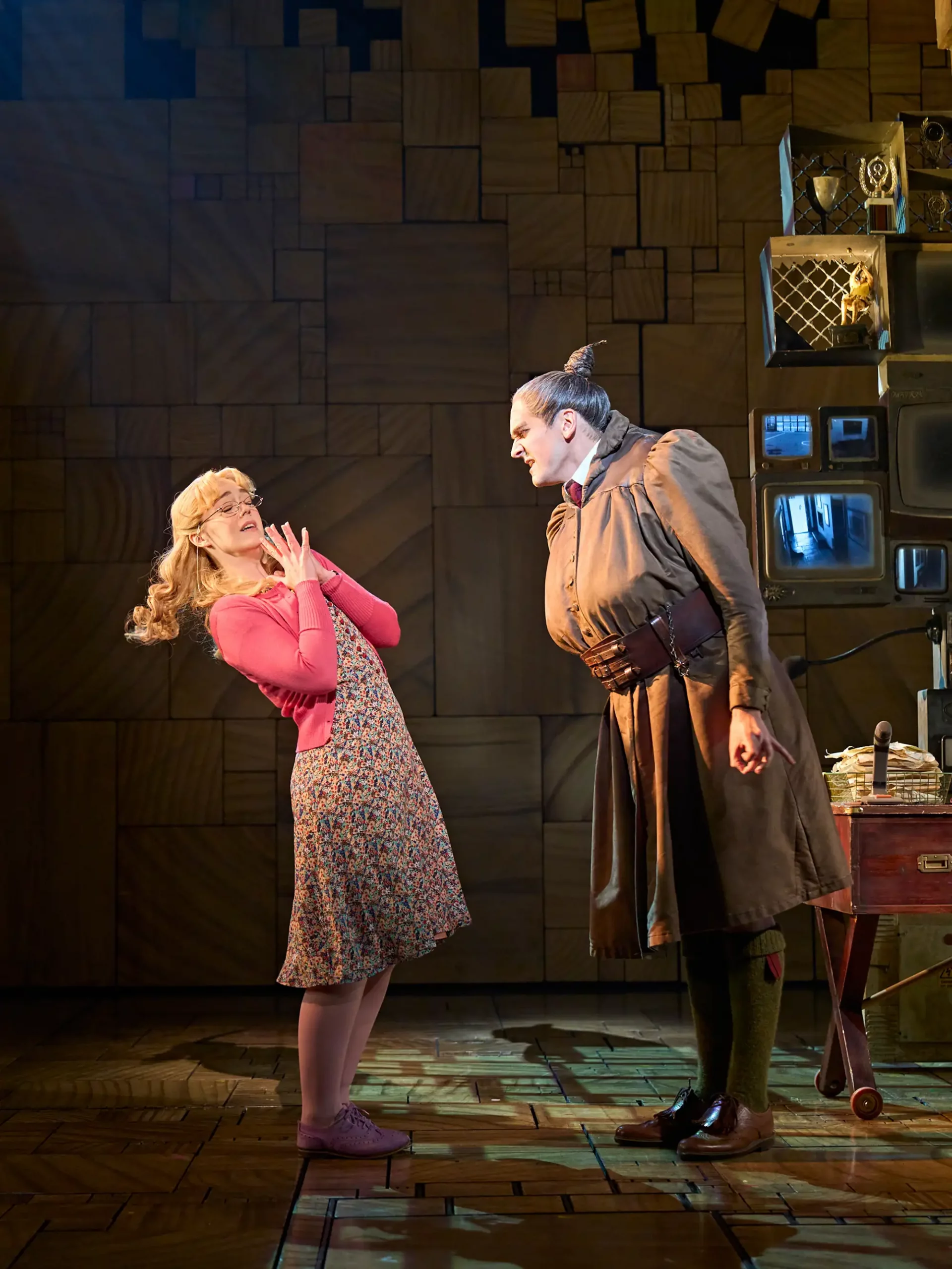 Szenenbild aus dem Matilda Musical mit Schulleiterin in drohender und Lehrerin in unterwürfiger Haltung und im Hintergrund im Regal Pokalen und Bildschirm