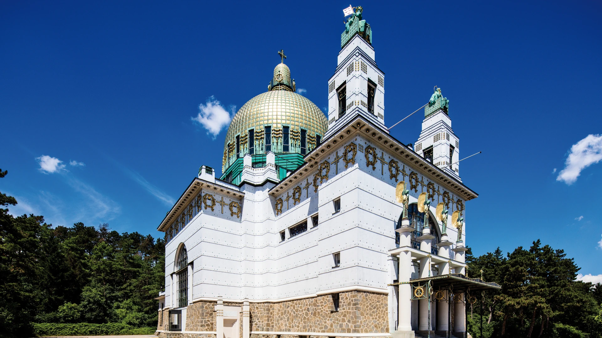 Dieses Foto zeigt die Otto Wagner Kirche am Steinhof in Wien, ein Meisterwerk des Jugendstils. Die markante goldene Kuppel und die kunstvoll verzierten Türme heben sich eindrucksvoll vor dem tiefblauen Himmel ab. Die weiße Fassade mit goldenen Elementen verleiht dem Bauwerk eine elegante und harmonische Ausstrahlung.