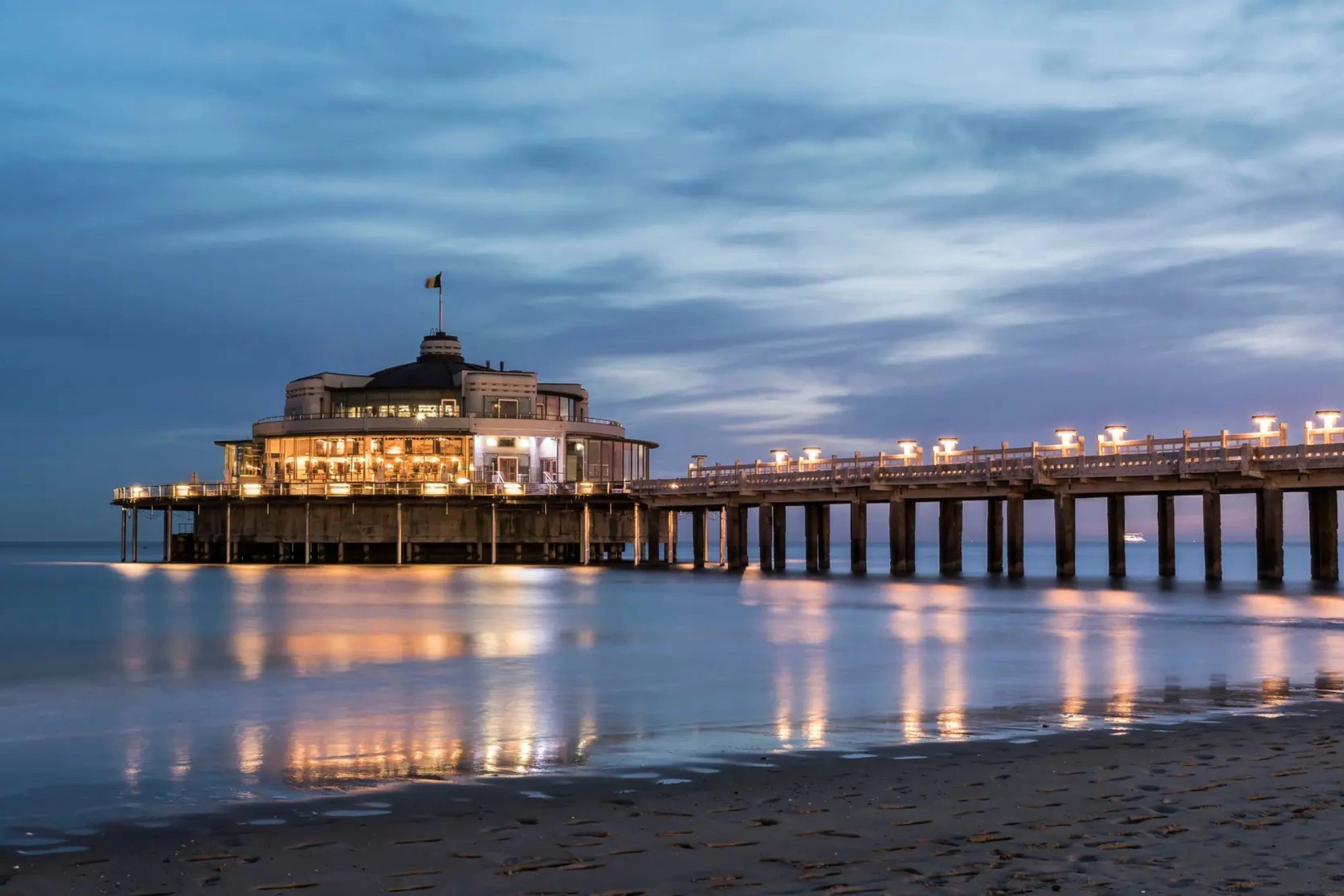 Beleuchteter belgischer Pier in der Abenddämmerung, der warmes Licht ausstrahlt und eine einladende Atmosphäre am Wasser schafft, umgeben von sanften Wellen und dem schwindenden Tageslicht.