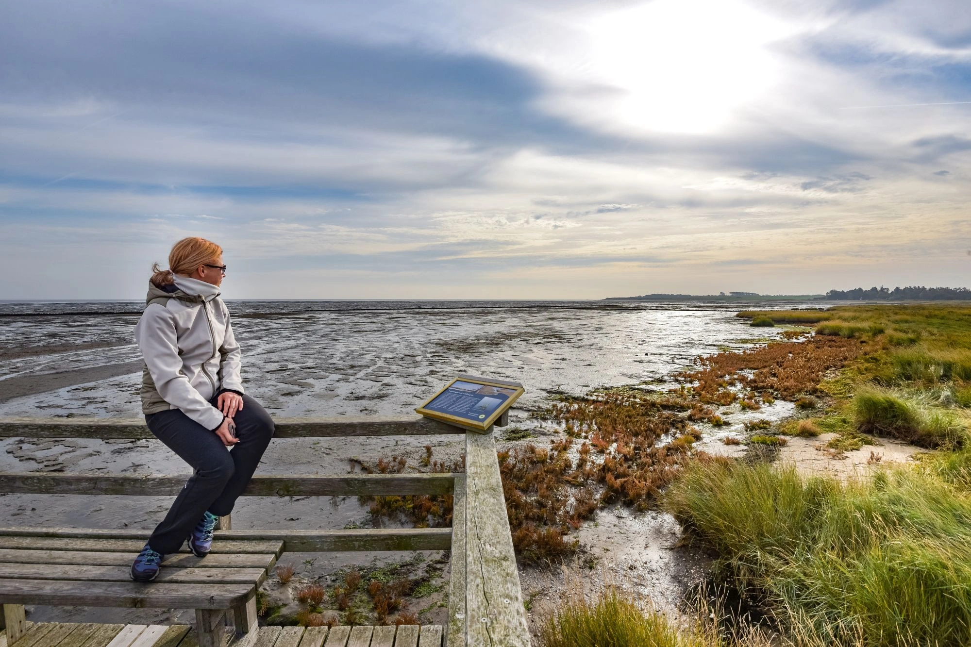 Person mit hellgrauer Jacke sitzt auf einer Holzbank an einem Pfahl mit Infotafel, Blick auf Wattlandschaft und Himmel mit Sonne.