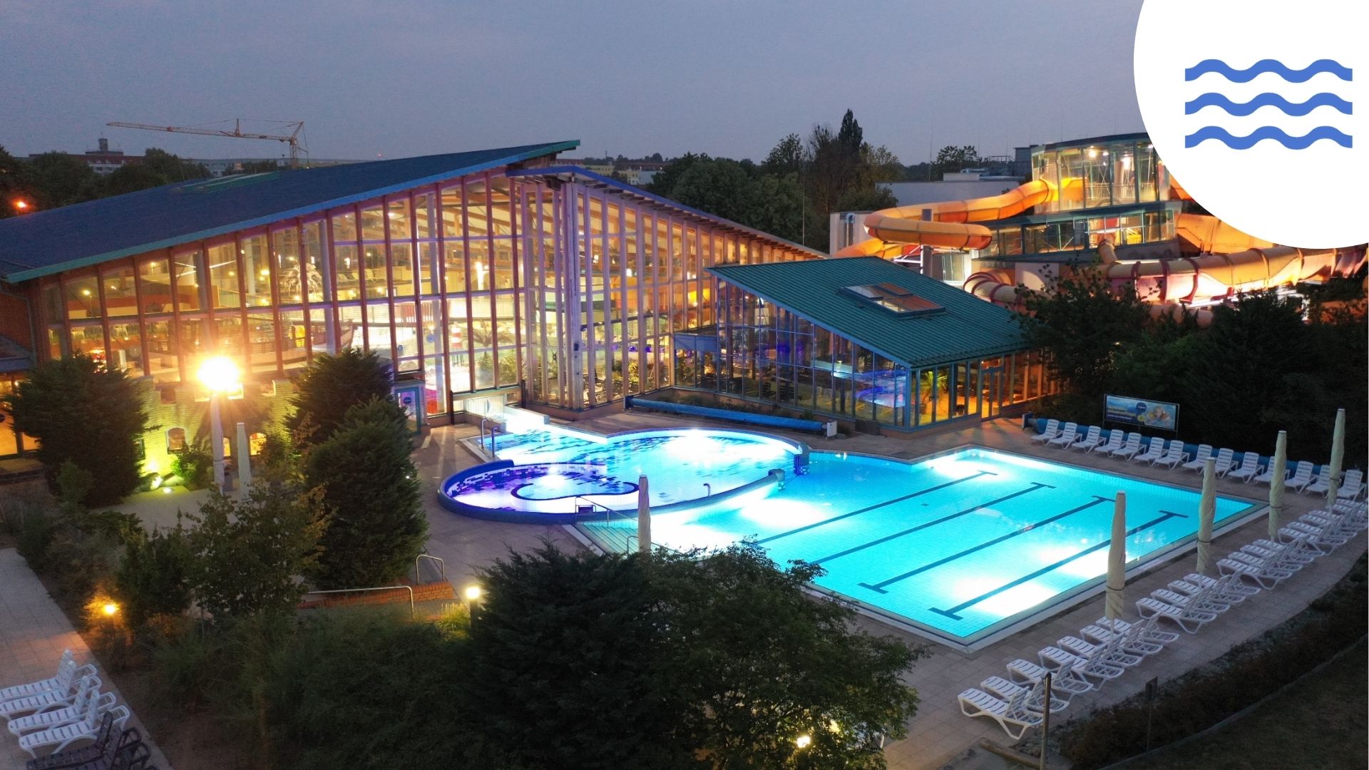 Wonnemar Resort Wismar Außenansicht am Abend von erhöhter Position aus über den Pool. Hinter dem Pool steht die große Schwimmhalle mit kompletter Glasfassade. Im Hintergrund sieht man einen Glasturm, aus dem Wasserrutschen nach unten zurück in die Haupthalle führen. Schwimmbad, Pool und Außenbereich sind beleuchtet.