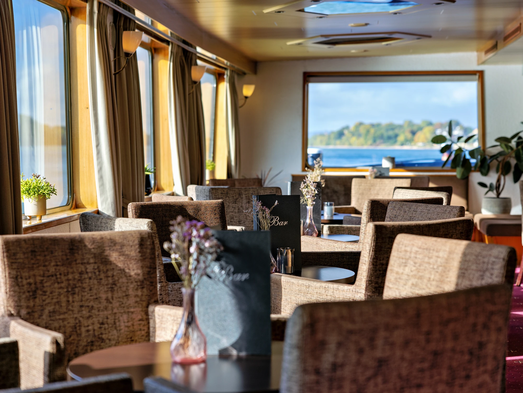 Bar der MS Olympia im Sonnenlicht mit gemütlichen Stühlen und kleinen runden Tischen vor Fenstern