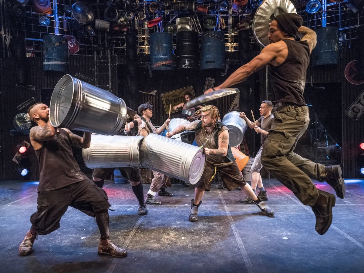 Energiegeladene Bühnenperformance der Rhythmus-Show STOMP 2025: Die Künstler nutzen Metallmülltonnen und Deckel als Percussion-Instrumente, während sie in einer synchronen Choreografie interagieren. Die industrielle Kulisse mit aufgehängten Fässern und Schildern verstärkt die rohe, urbane Ästhetik der Inszenierung.