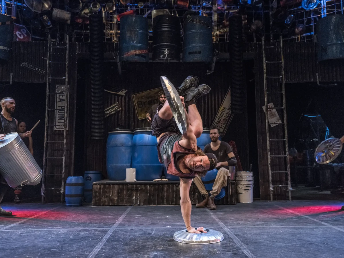 Ein Künstler von STOMP 2025 zeigt eine beeindruckende akrobatische Einlage, während er auf einem Mülleimerdeckel balanciert. Die Performance kombiniert Rhythmus, Tanz und körperliche Geschicklichkeit zu einem mitreißenden Bühnenerlebnis.