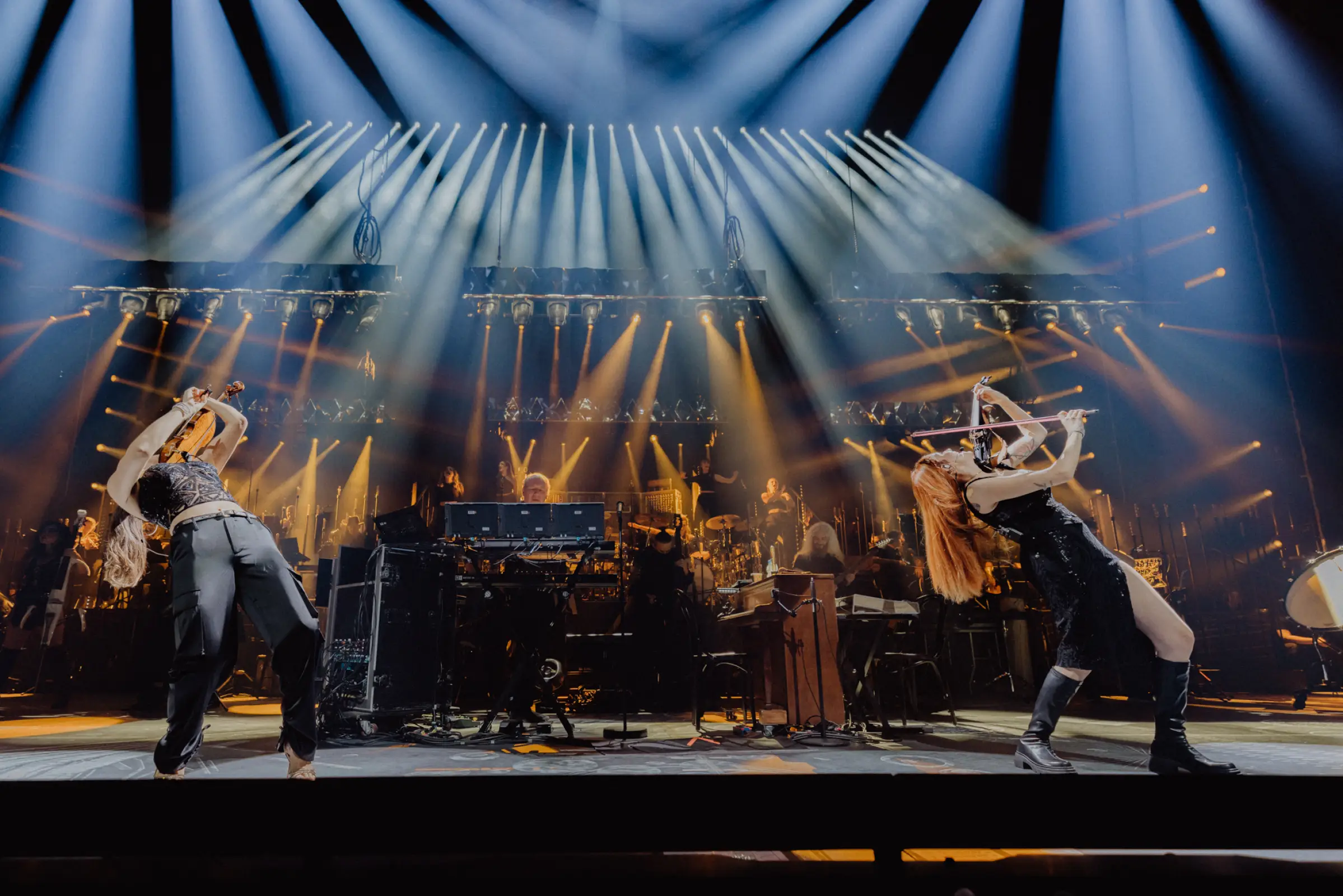 Zwei Musikerinnen performen leidenschaftlich auf der Bühne während der Hans Zimmer Show, begleitet von intensiver Beleuchtung und einem Orchester im Hintergrund. Die dynamische Körperhaltung und die Lichteffekte verstärken die kraftvolle Atmosphäre des musikalischen Erlebnisses.