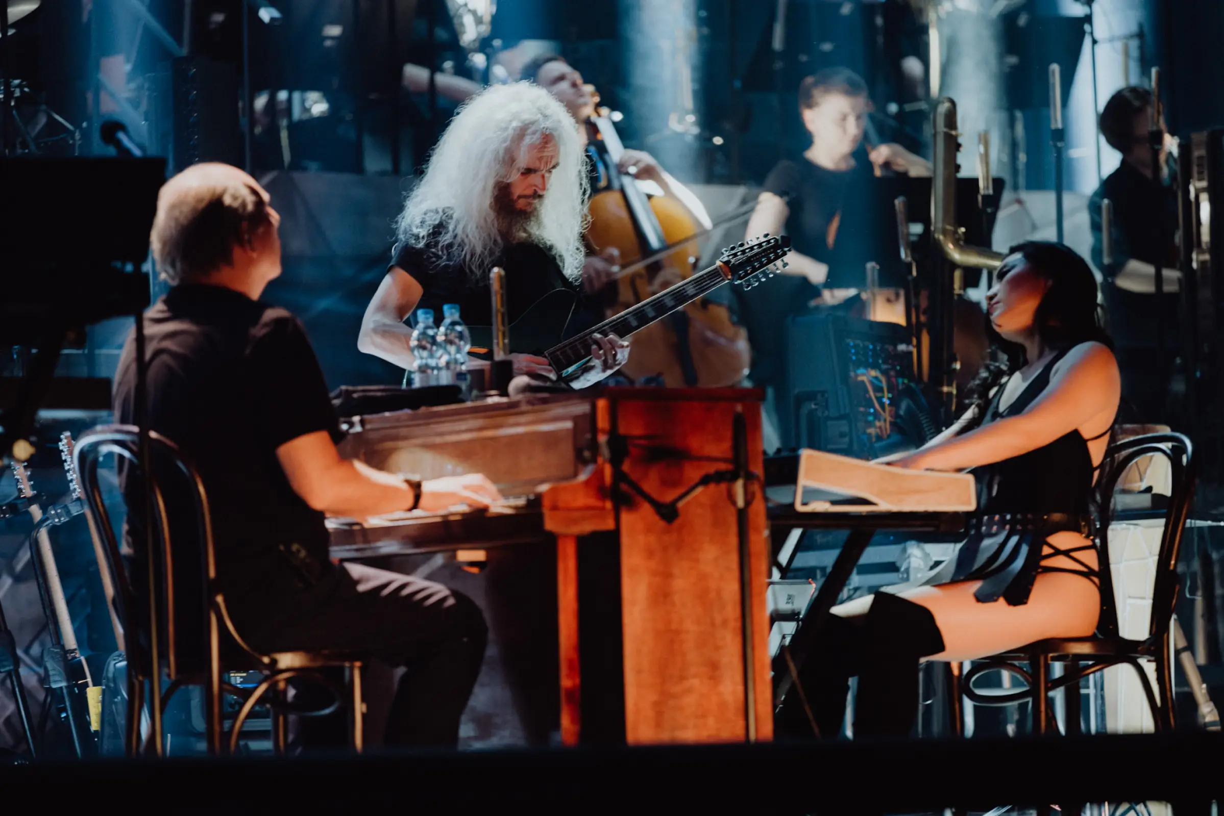 Eine intime Szene auf der Bühne, bei der Hans Zimmer und seine Mitmusiker in tiefem Fokus musizieren. Ein Gitarrist mit langen, weißen Haaren und eine Keyboard-Spielerin verleihen der Performance eine besondere Intensität. Die Beleuchtung schafft eine stimmungsvolle Atmosphäre und betont die musikalische Hingabe der Künstler.