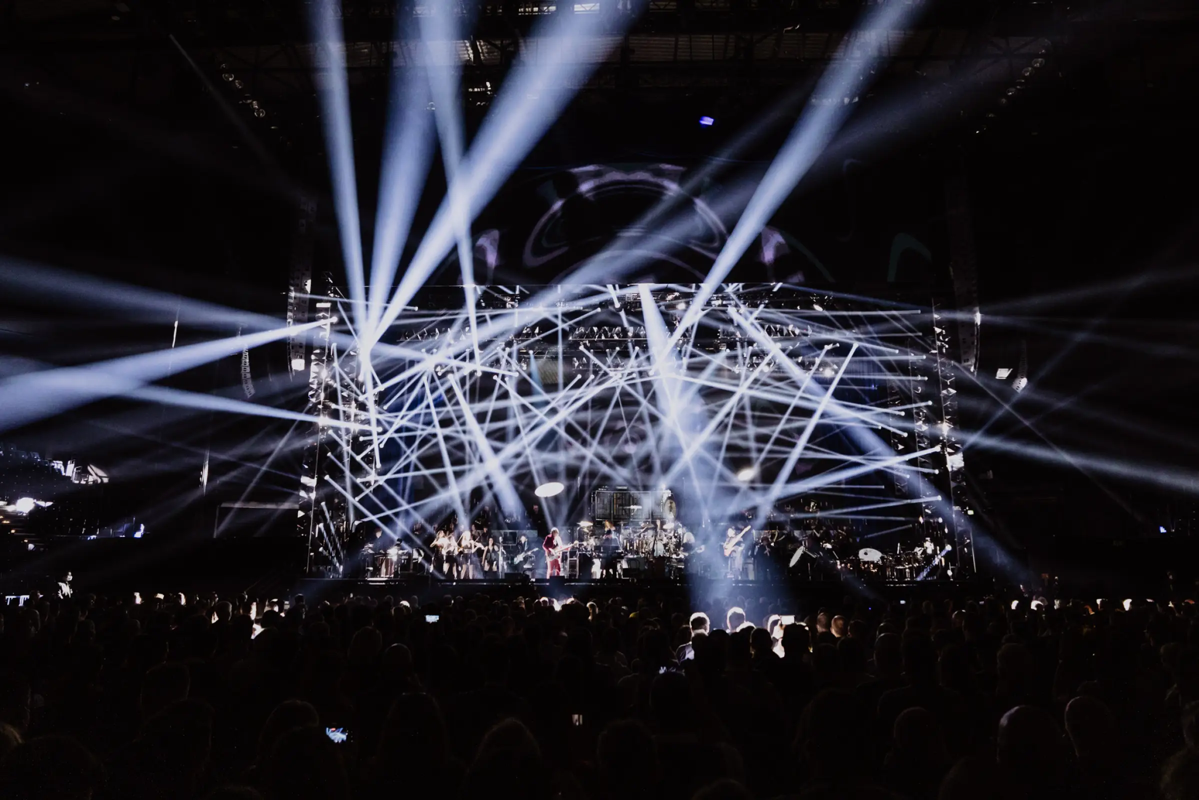 Hans Zimmer Show mit intensiven, kreuzenden weißen Lichtstrahlen, die die Bühne durchziehen. Die Musiker und Instrumente sind im Hintergrund sichtbar, während das Publikum in den Vordergrund rückt und die eindrucksvolle Lichtinszenierung die Szene dominiert.
