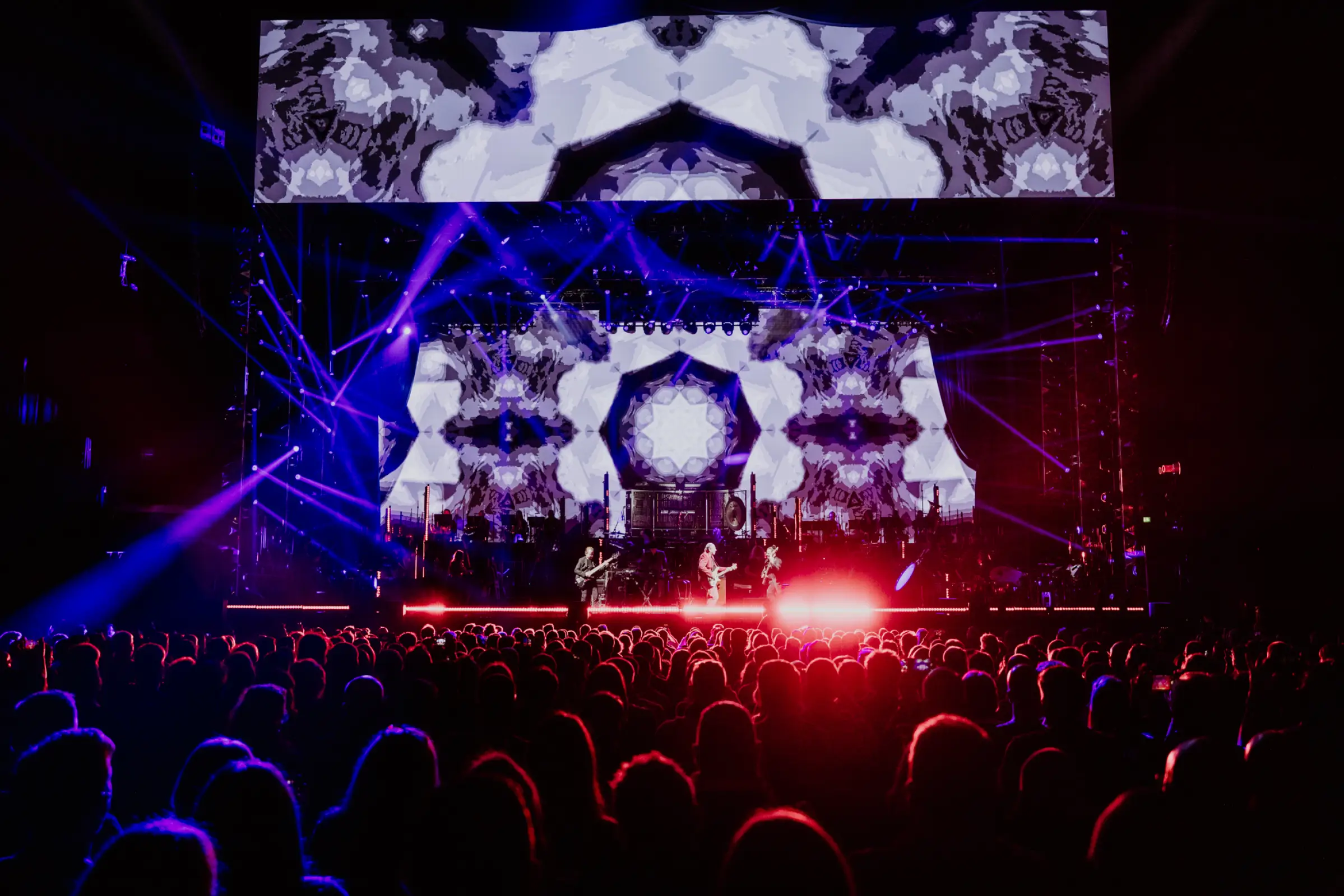 Hans Zimmer Show mit beeindruckender visueller Projektion, die kaleidoskopische Muster in Schwarz-Weiß auf großen Bildschirmen zeigt. Die Bühne wird von blauen und roten Lichtstrahlen durchzogen, während die Silhouetten des Publikums im Vordergrund sichtbar sind.