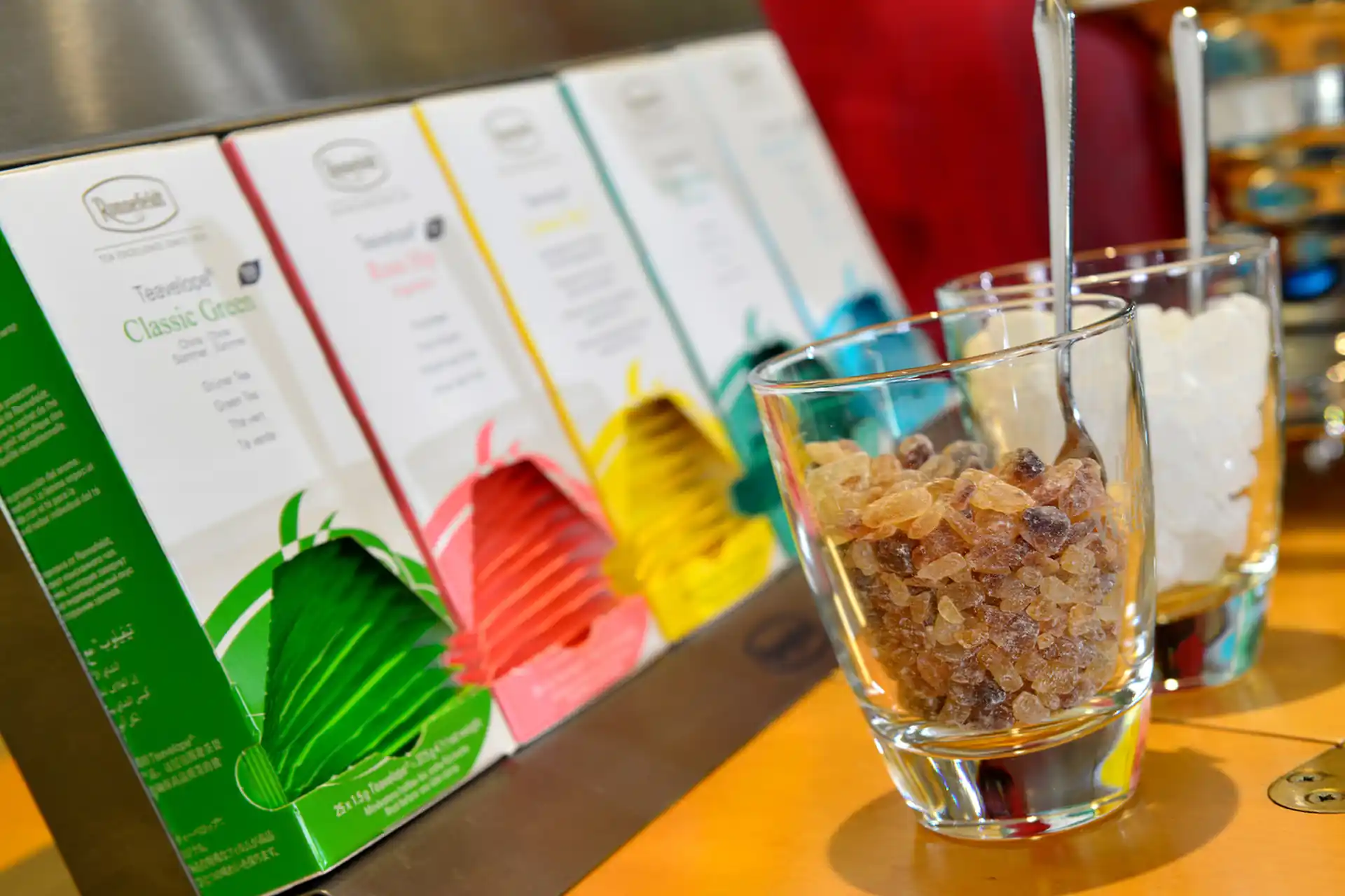 Glas mit braunem Kandiszucker und Glas mit weißem Kandiszucker vor aufgestellten Teeverpackungen in verschiedenen Farben