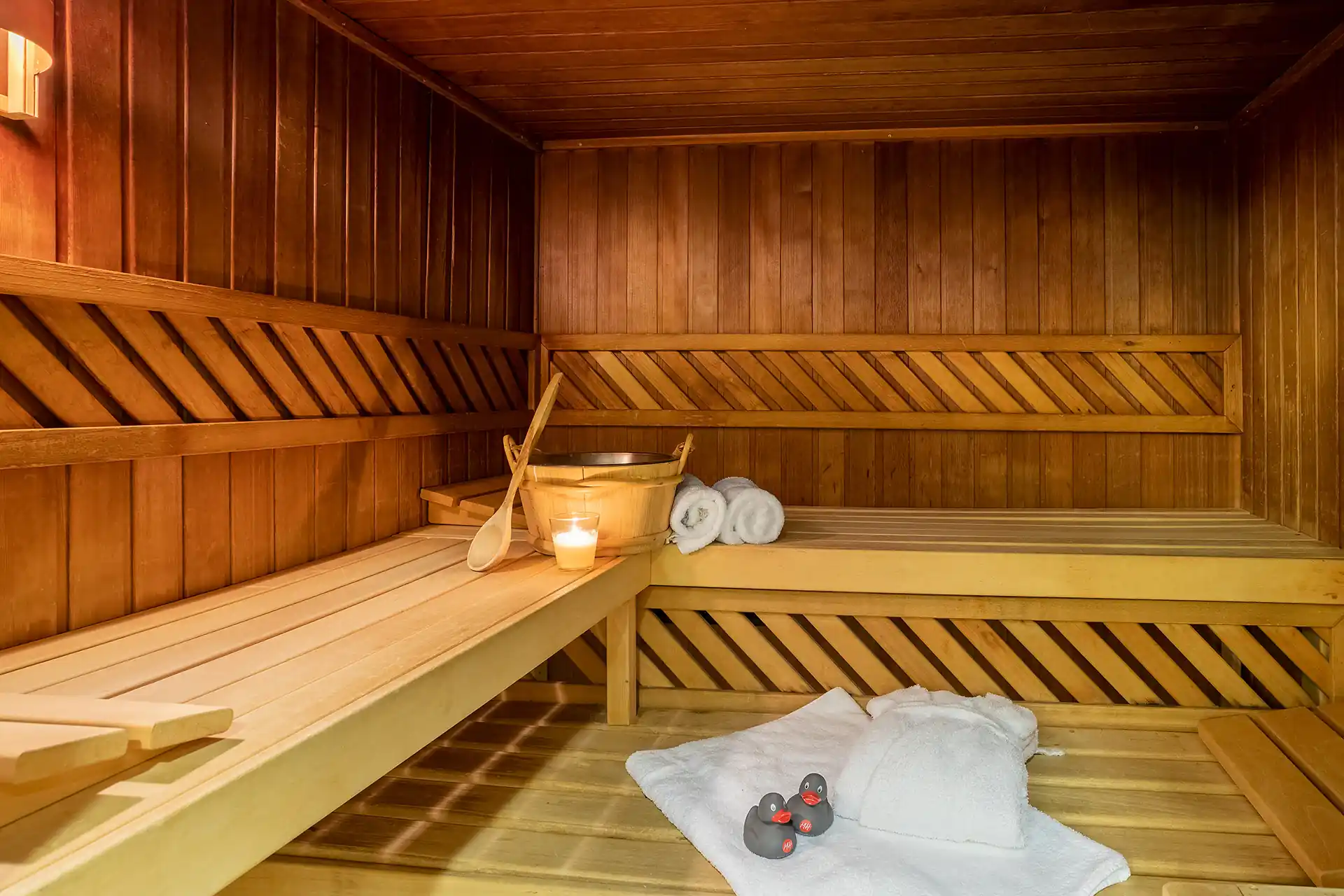 Saunaraum mit Holzbänken, Holzwänden, einem Holzeimer mit Kelle, einer brennenden Kerze, zwei zusammengerollten Handtüchern auf der Bank und einem weißen Bademantel mit drei Gummienten auf dem Boden