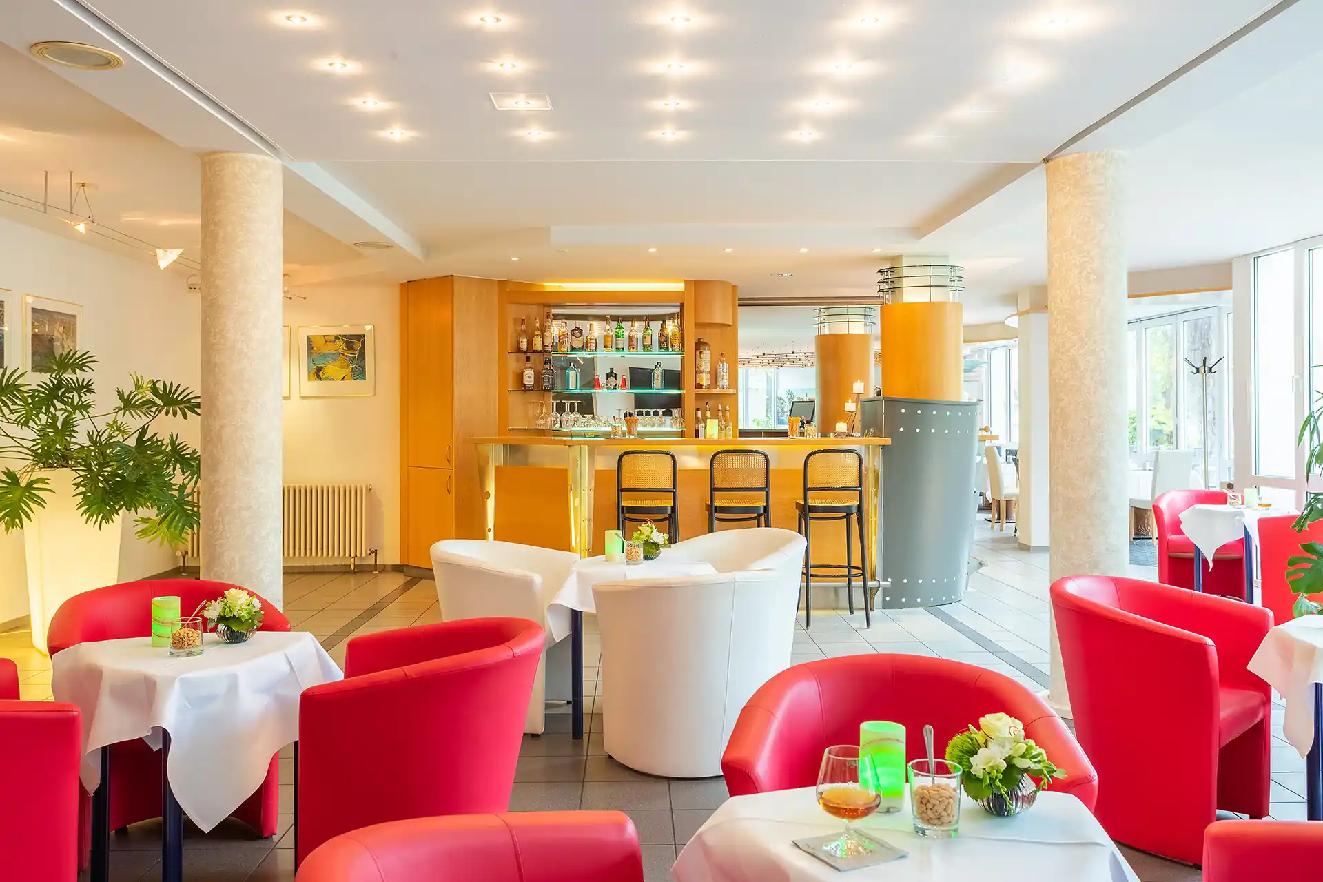 Lobby des ACHAT Hotels Suhl mit roten und weißen Sesseln, runden Tischen mit weißen Tischdecken, einer Bar mit Hockern und großen Fenstern im Hintergrund