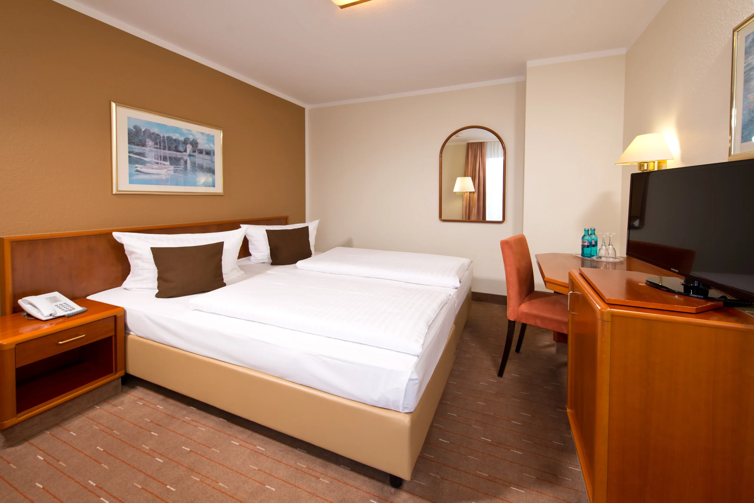 Hotelzimmer mit Doppelbett, Nachttisch mit Telefon, Schreibtisch mit Stuhl, Flachbildfernseher, Spiegel und Wandbild