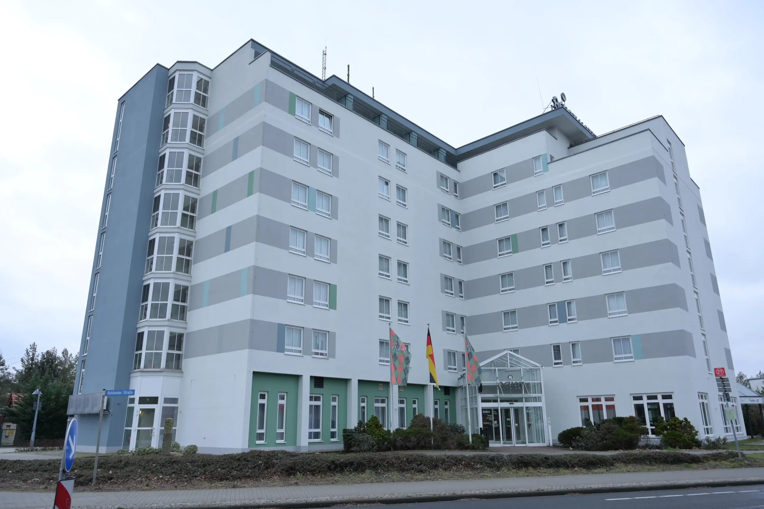 Mehrstöckiges Hotelgebäude mit weißer Fassade und grauen horizontalen Streifen, Eingangsbereich mit Glasvordach und mehreren Flaggen davor