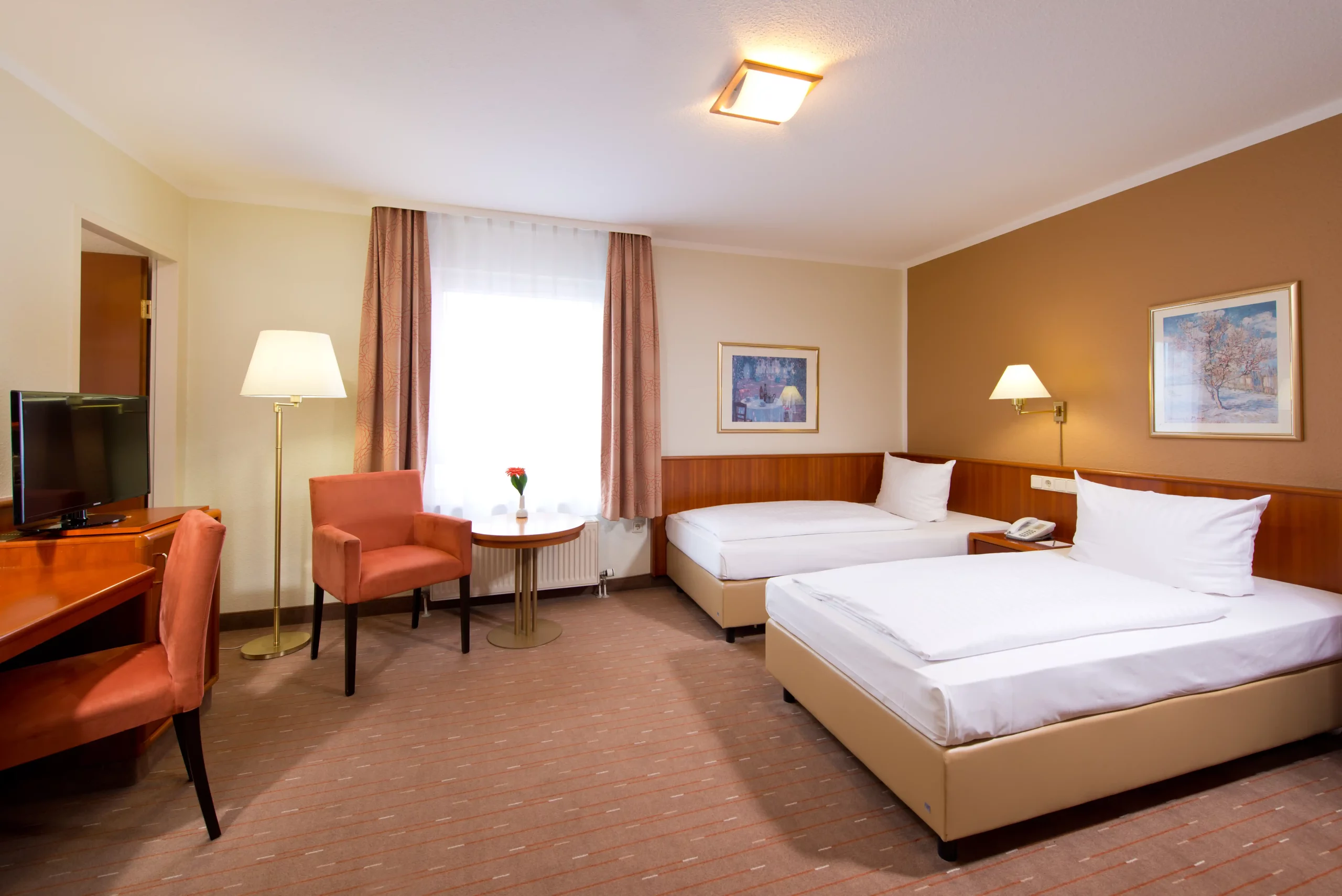 Hotelzimmer mit zwei Einzelbetten, einem kleinen runden Tisch mit zwei Sesseln, einem Schreibtisch mit Stuhl, einem Fernseher und zwei Wandbildern.