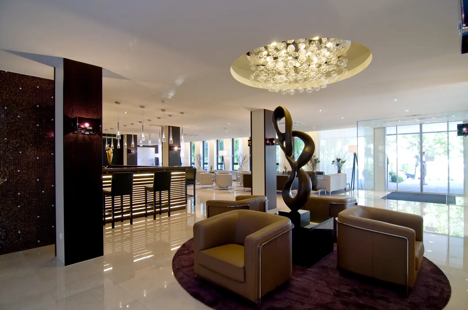 Achat Hotel Müchen Süd Lobby mit hellen Fliesen. Ledersessel stehen um eine abstrakte Brozeskulptur herum. Helles Tageslicht fällt durch die bodentiefen Fenster und die Eingangstür ins Gebäude. Im Hintergrund sind eine Bar mit hohen Stühlen und weitere Sitzgelegenheiten zu sehen.