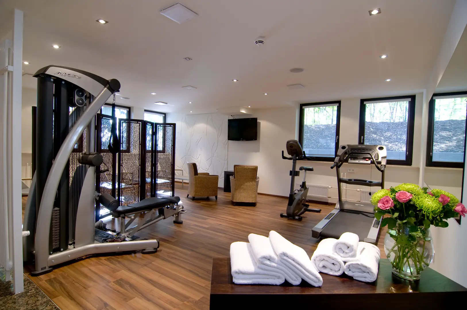 Achat Hotel München Süd Fitnessbereich mit Laufband, Indoor Bike und Gewichten. Weiter hinten stehen Sessel vor einem Fernseher an der Wand. Im Vordergrund liegen frische Handtücher auf einer Anrichte mit frischen Schnittblumen.