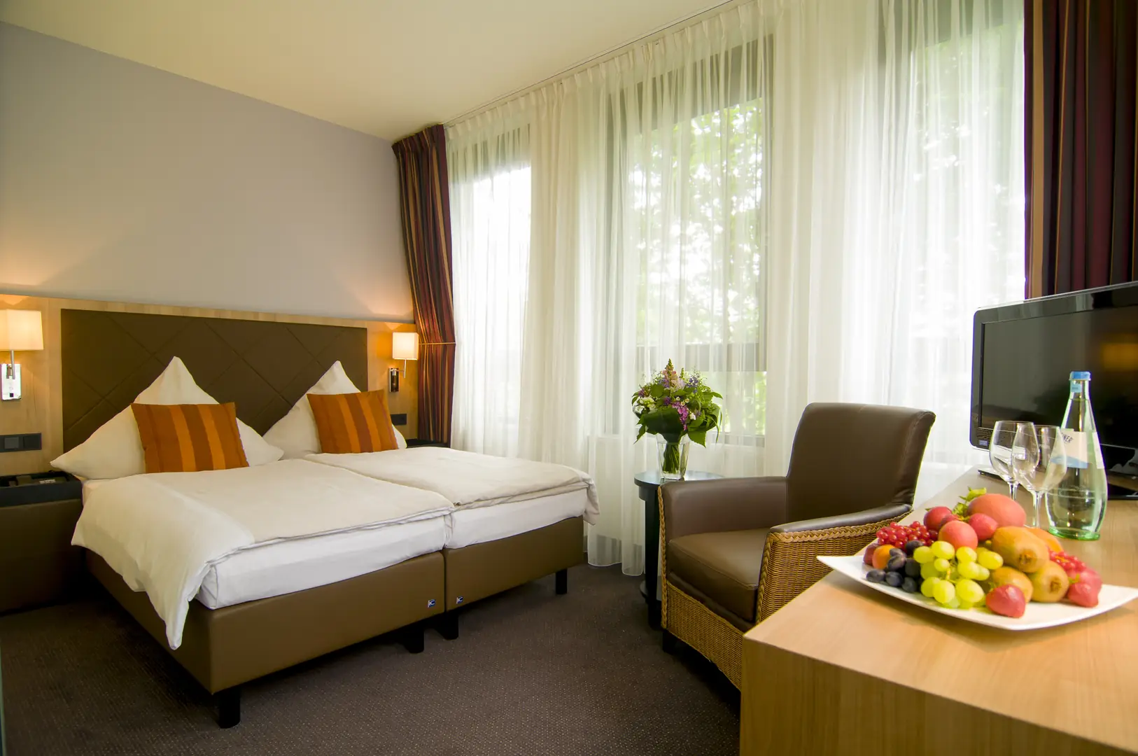 Achat Hotel München Süd Gästezimmer mit Doppelbett, Sessel und Fernseher. Im Vordergrund steht ein frischer Obstkorb und eine Flasche Mineralwasser mit Gläsern. Durch die Vorhänge fällt warmes Tageslicht ins Zimmer. Neben dem Sessel steht ein Strauß frischer Schnittblumen.
