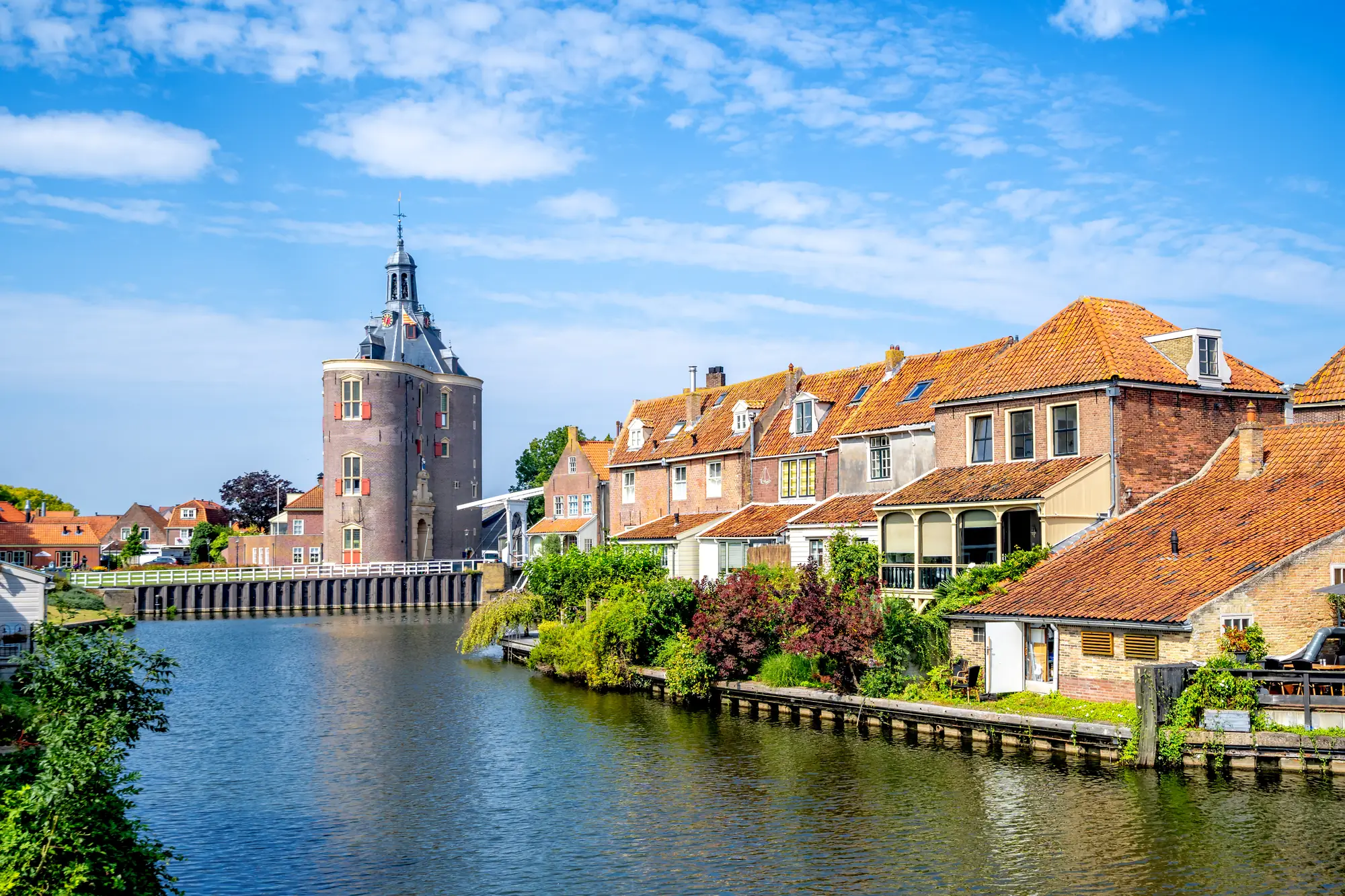 Fluss mit historischen Backsteinhäusern und einem runden Turm mit spitzem Dach unter blauem Himmel