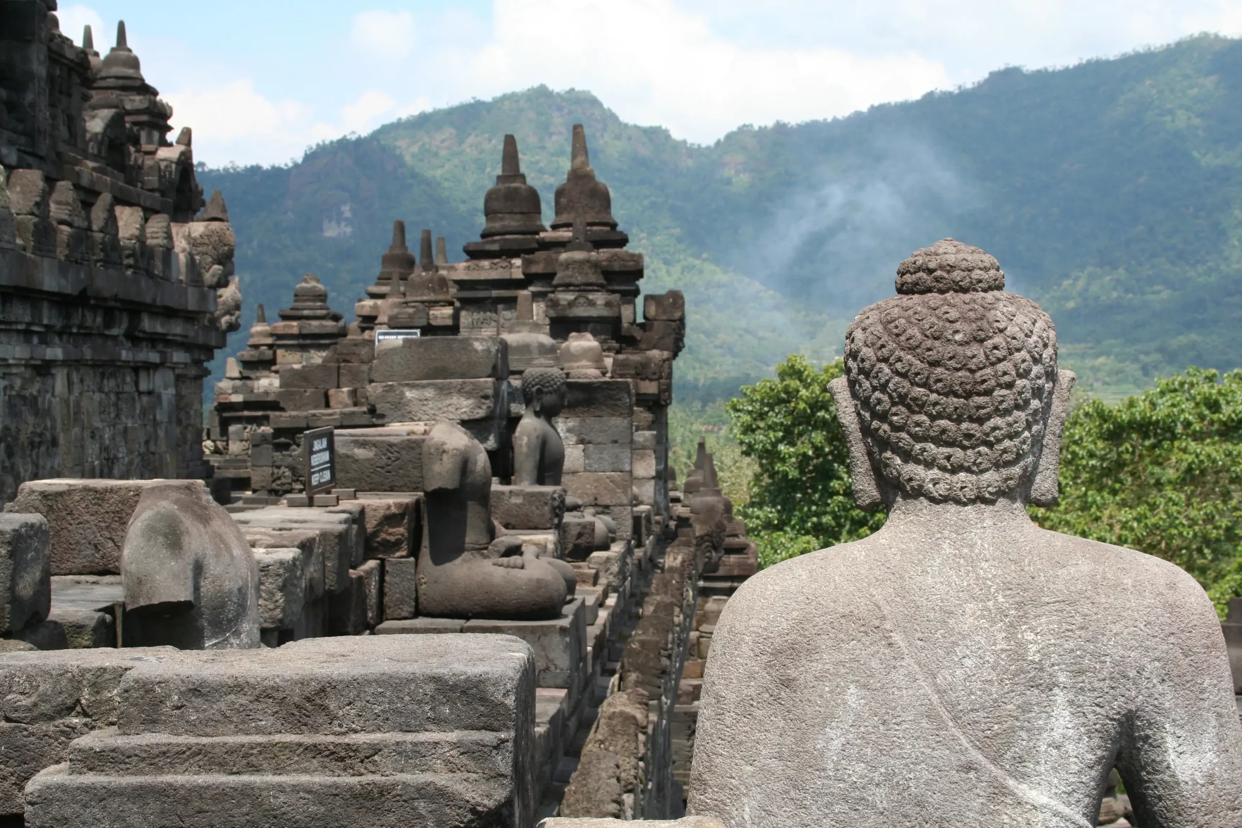Rückansicht einer großen Buddha-Statue aus Stein vor einem antiken Tempel mit weiteren Buddha-Statuen und Bergen im Hintergrund in Java