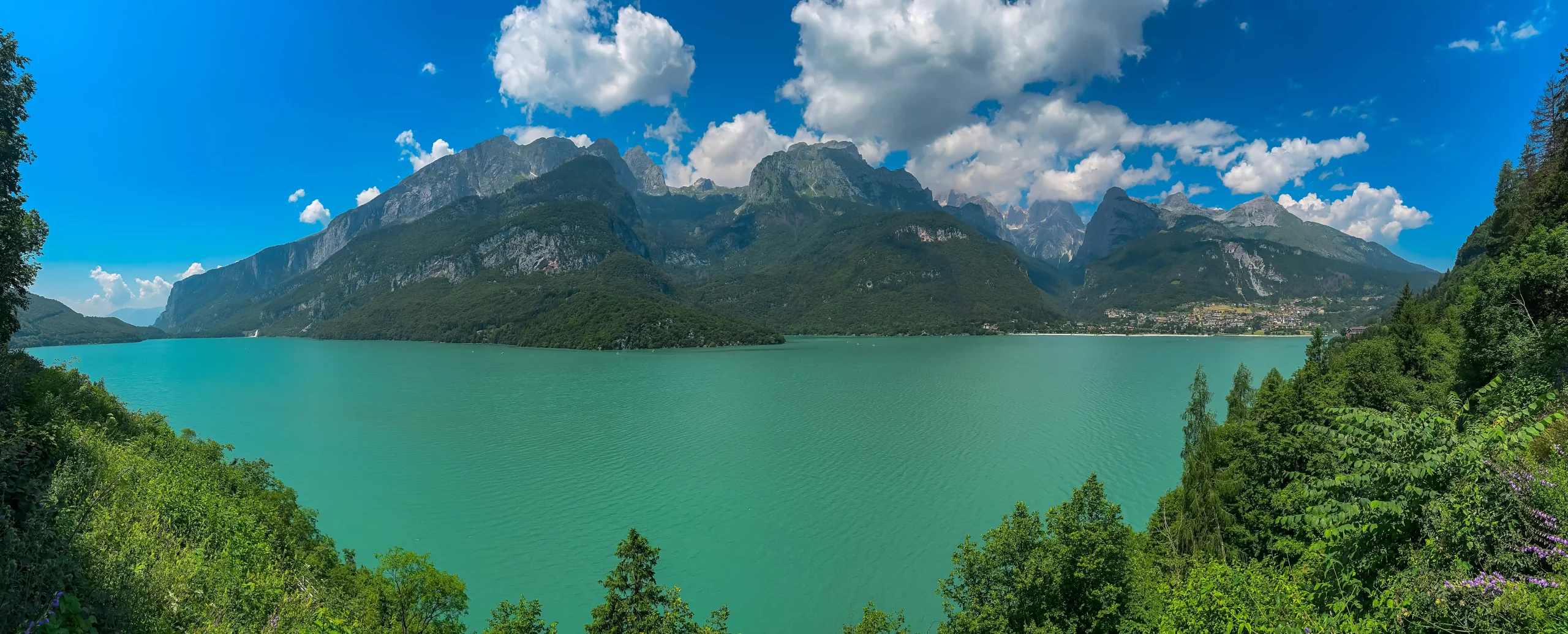 Greenery frames alpine retreat village at idyllic mountain lake Lago di Molveno with scenic view of majestic mountains Brenta Dolomites, National Park ... Mehr anzeigen