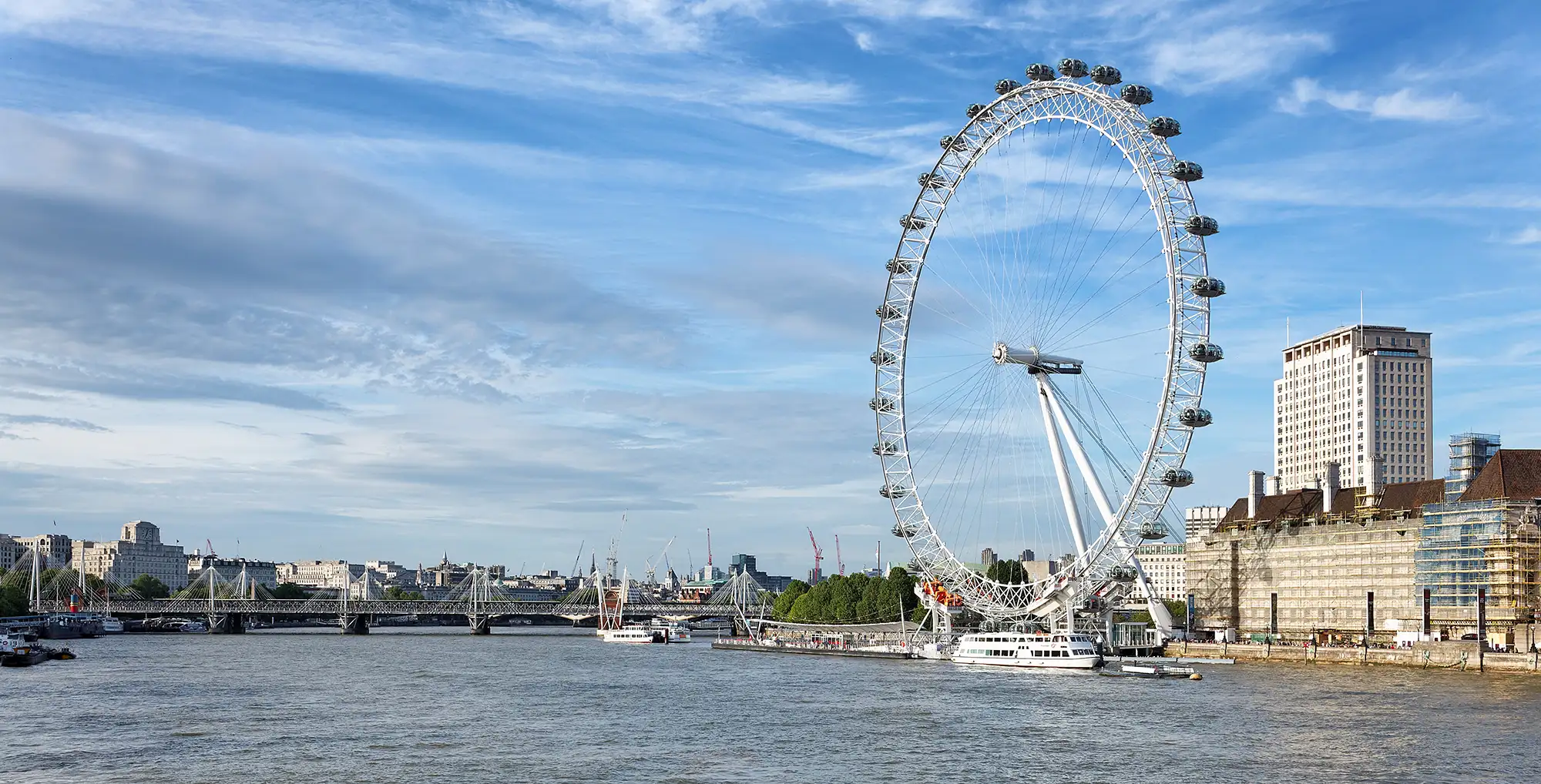 Das Bild zeigt das London Eye, eines der bekanntesten Wahrzeichen Londons, das sich direkt an der Themse befindet. Das riesige Riesenrad bietet Besuchern eine atemberaubende Aussicht auf die Stadt. Mit einer Höhe von 135 Metern ist es eines der höchsten Riesenräder Europas. Die zahlreichen Kabinen sind klar verglast, sodass Passagiere einen unverstellten Blick auf die umliegenden Sehenswürdigkeiten wie die Houses of Parliament und die Westminster Abbey haben können. Im Hintergrund sind auch einige Gebäude der Londoner Skyline zu sehen, die das moderne Stadtbild von London unterstreichen. Der Himmel ist klar und blau, was auf einen schönen Tag hindeutet.