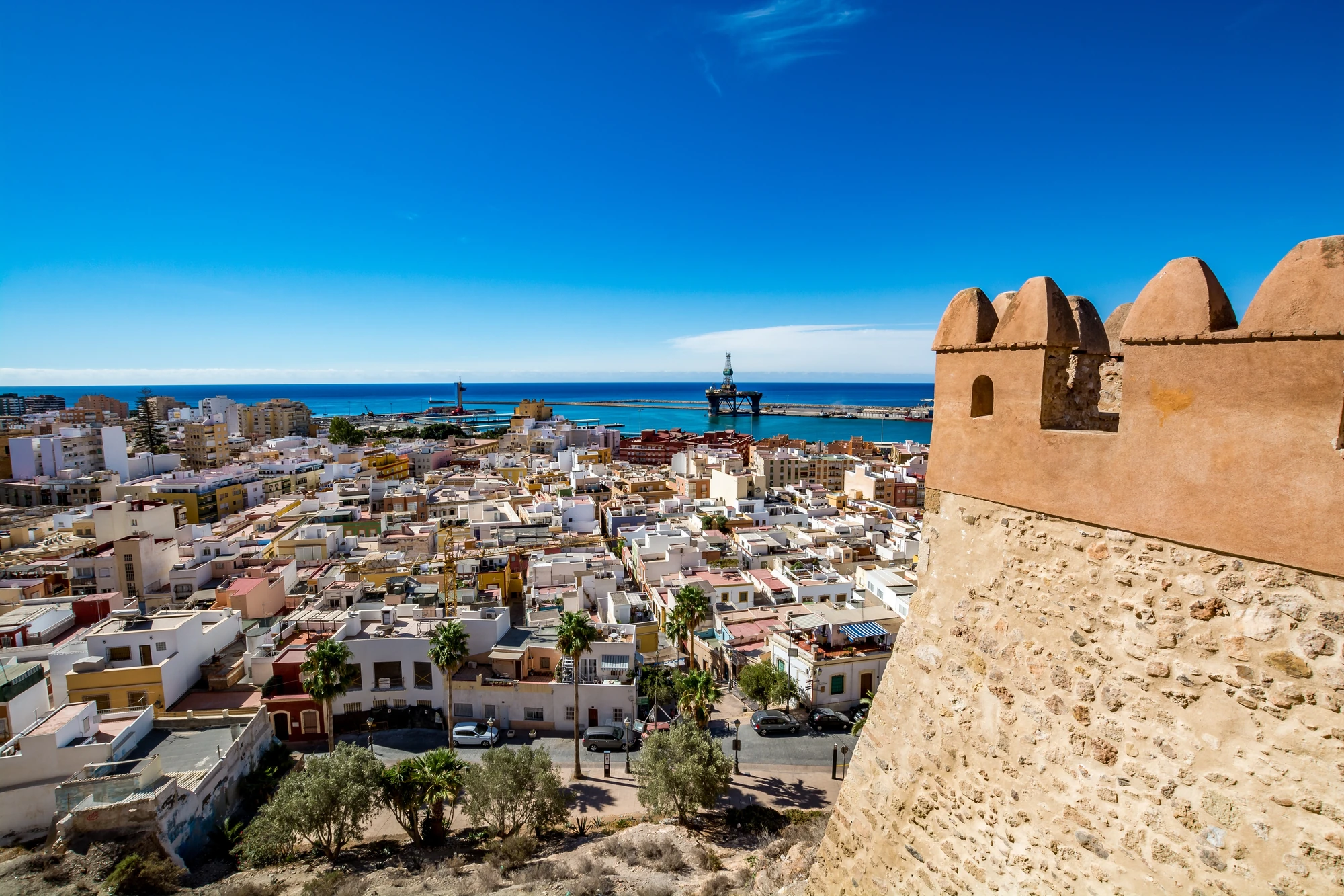 Almeria Blick von einer historischen Festungsmauer auf eine Stadt mit weißen Gebäuden und dahinter das blaue Meer unter klarem Himmel.